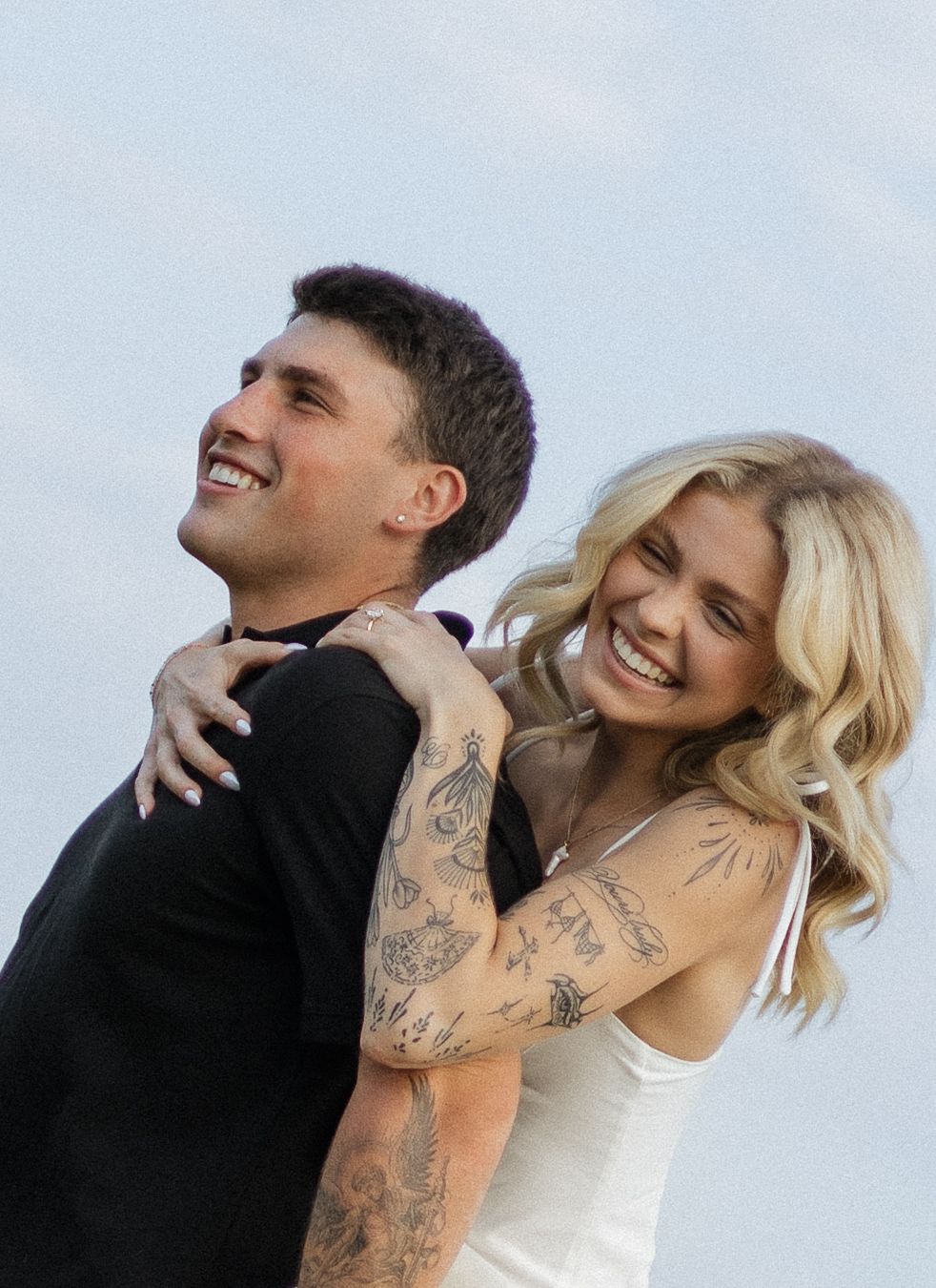 A laughing couple with tattoos embrace outdoors, the blonde woman in white hugging a dark-haired man in black from behind.