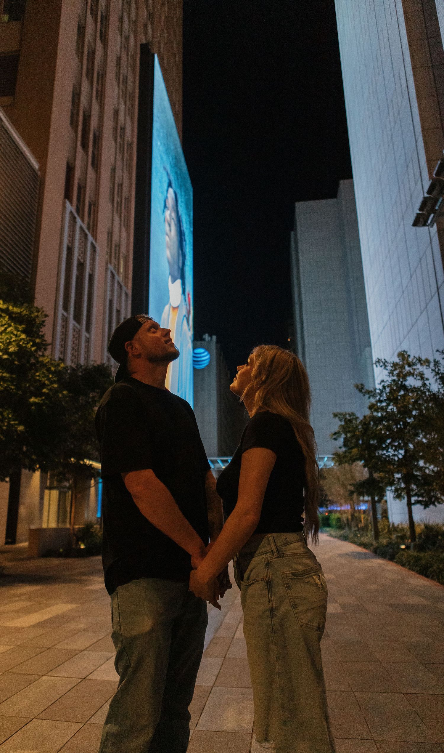A couple holds hands while gazing up at illuminated buildings in an urban setting at night. Dallas, Texas