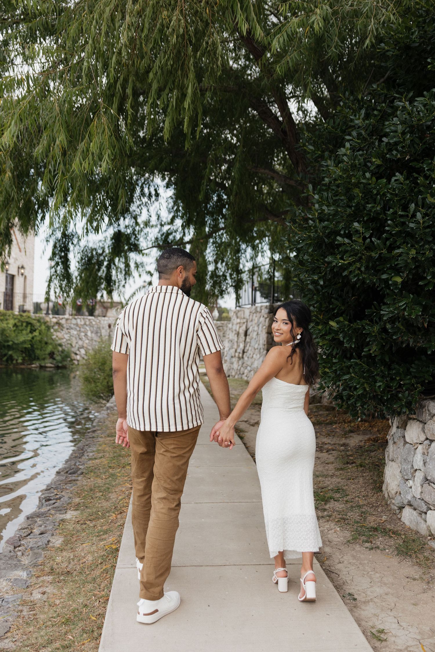Couple holding hands walking along a waterfront path, woman in white dress looking back, man in striped shirt.