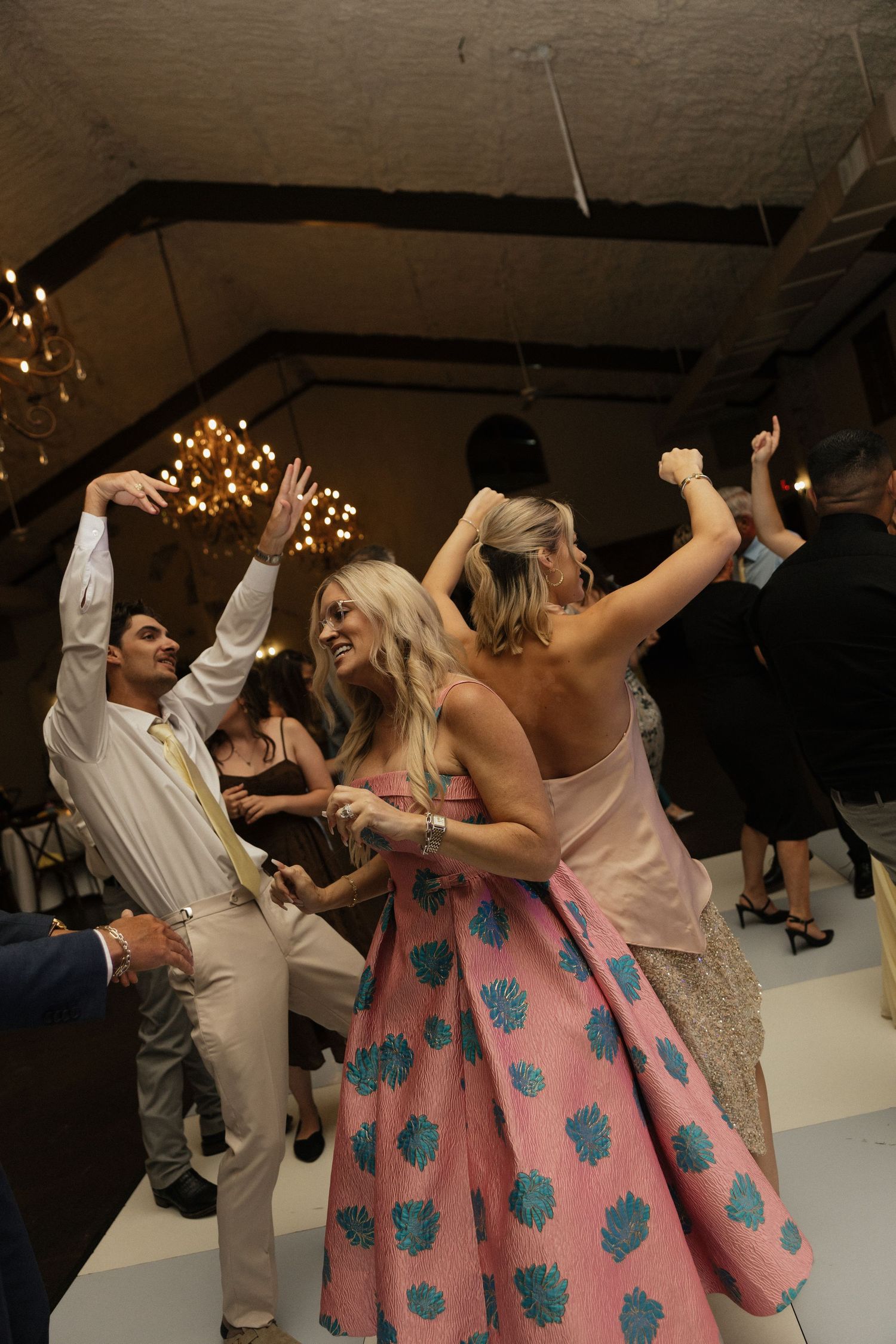 Guests celebrate at lively wedding reception as woman in pink floral dress dances with arms raised.