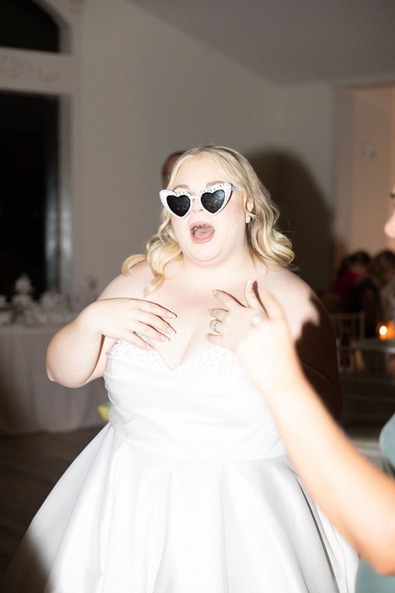 Blonde bride wearing heart-shaped sunglasses dancing joyfully at wedding reception in white gown.