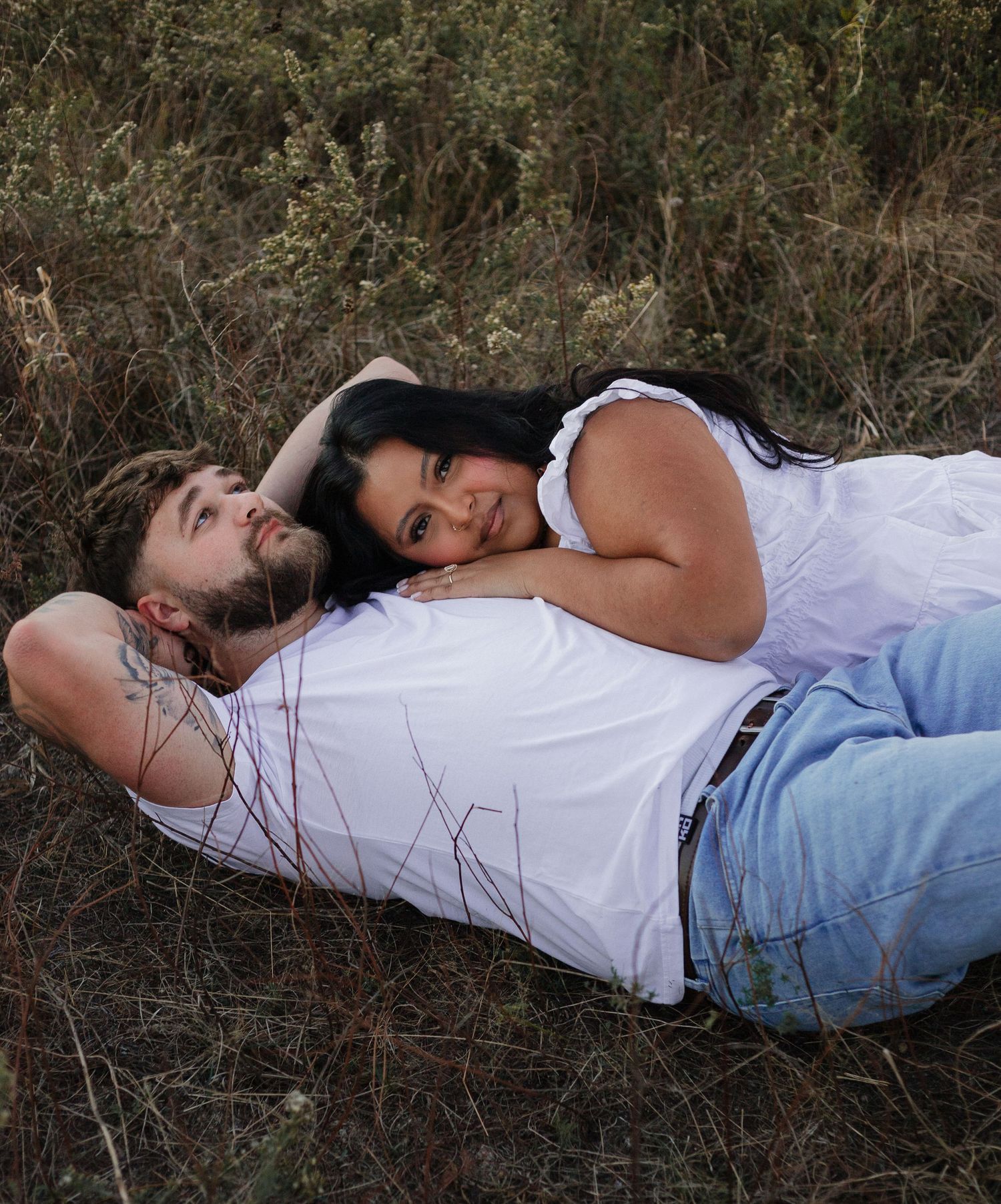 Couple relaxing together in grass field wearing matching white tops and light denim.