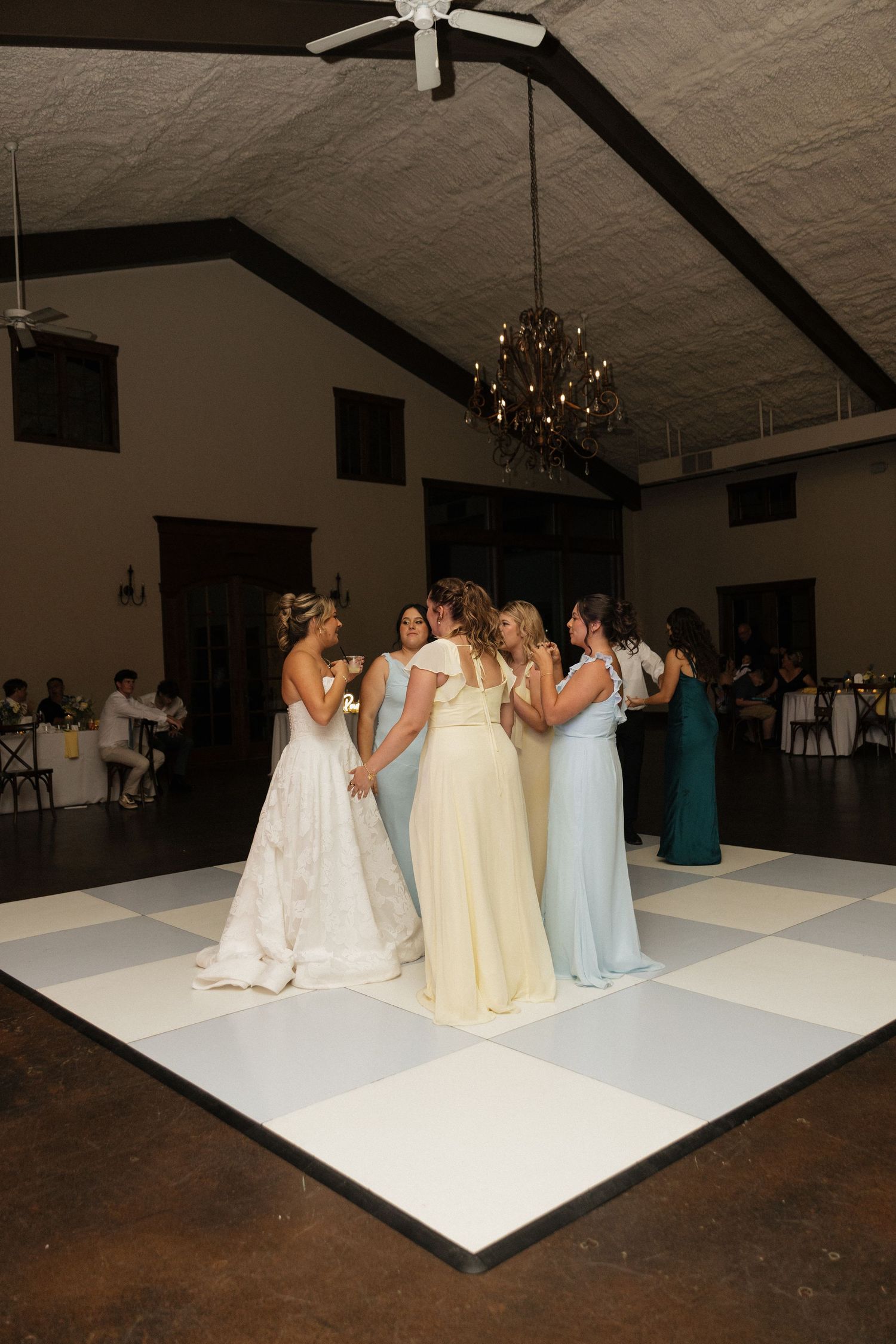 Bride and bridesmaids in pastel gowns share a tender group hug on a checkered dance floor at wedding reception.