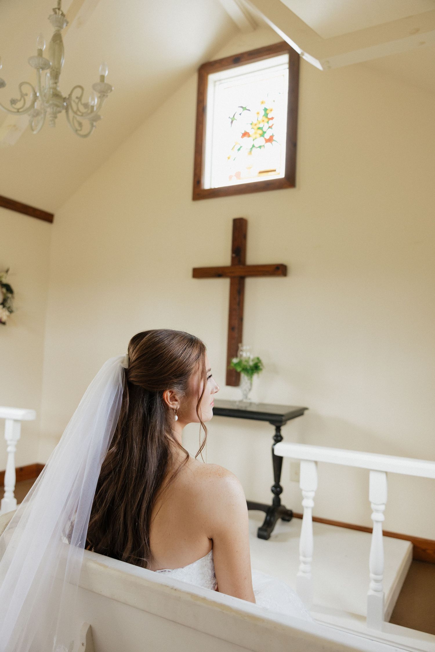 A bride in a white strapless gown and long veil sits in a church pew with a wooden cross and stained glass window visible.
