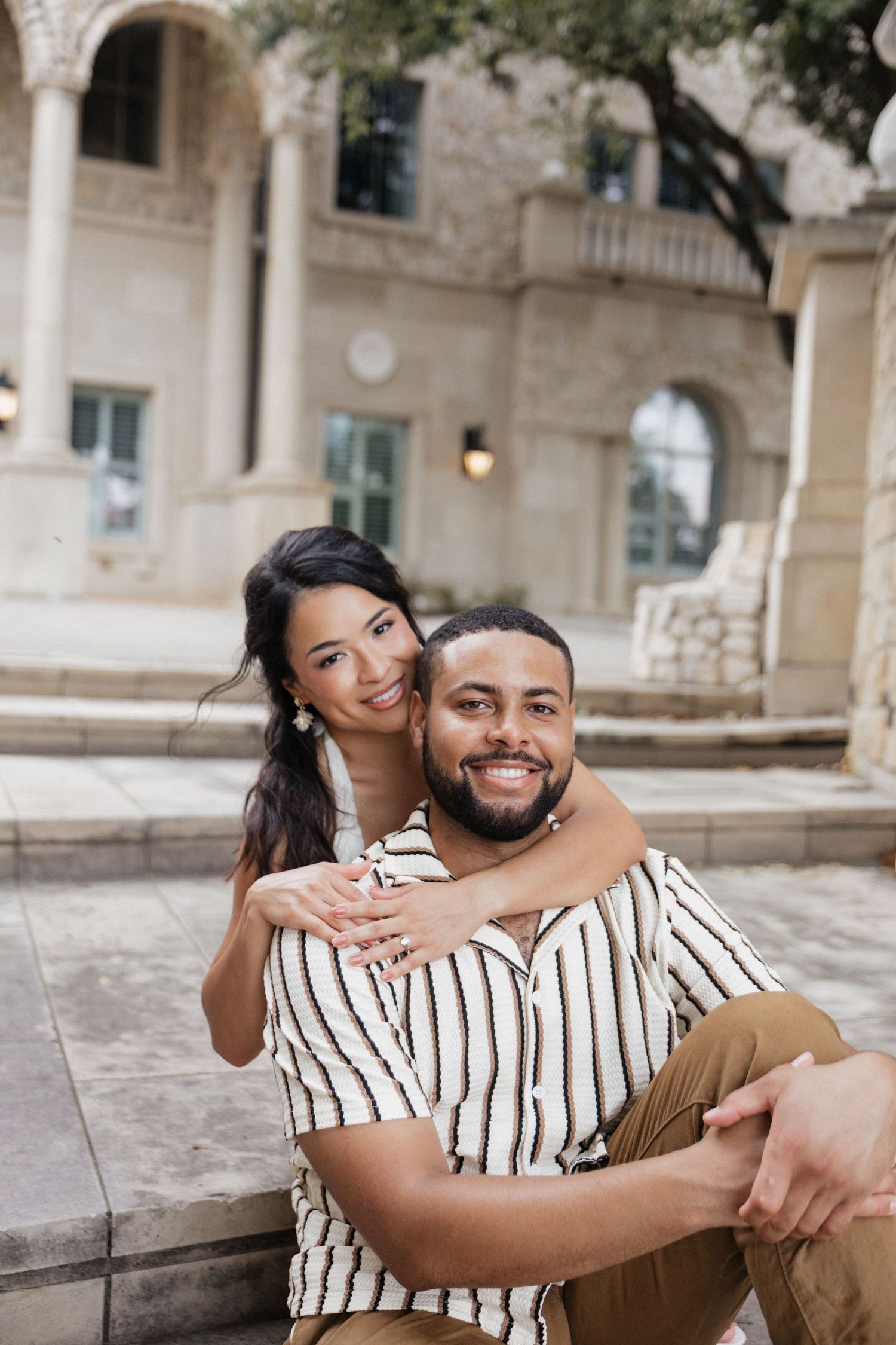 A smiling couple poses outdoors near a stone building, with the woman embracing the man from behind on steps.