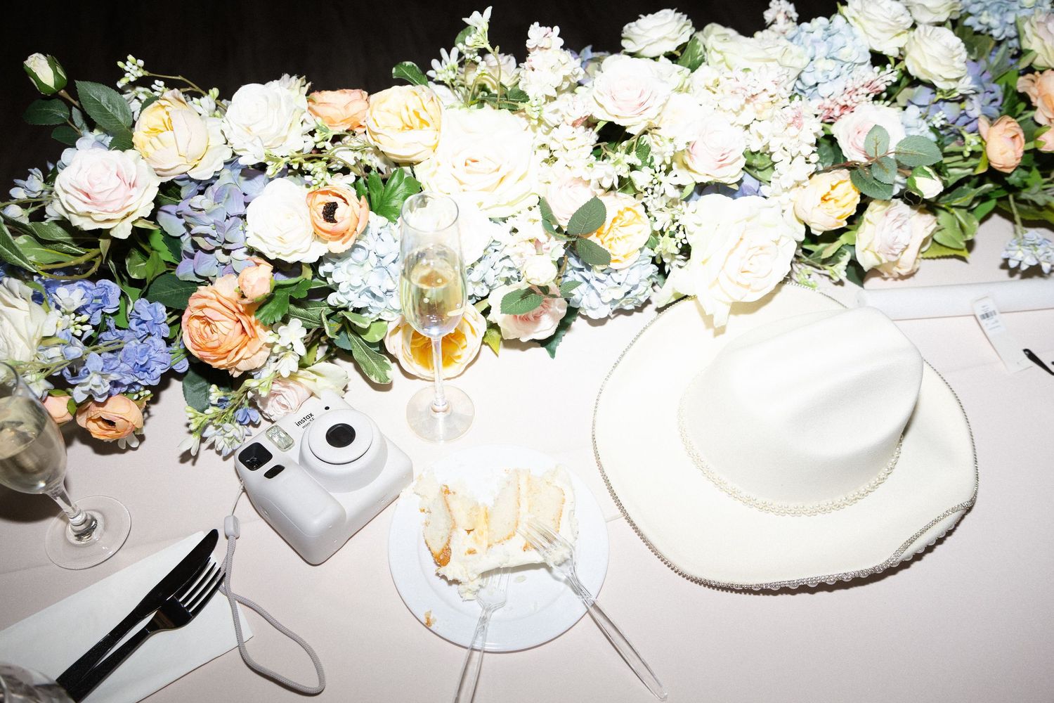 Wedding sweetheart table with pastel floral arrangement, white cowboy hat, Instax camera, and champagne glasses.