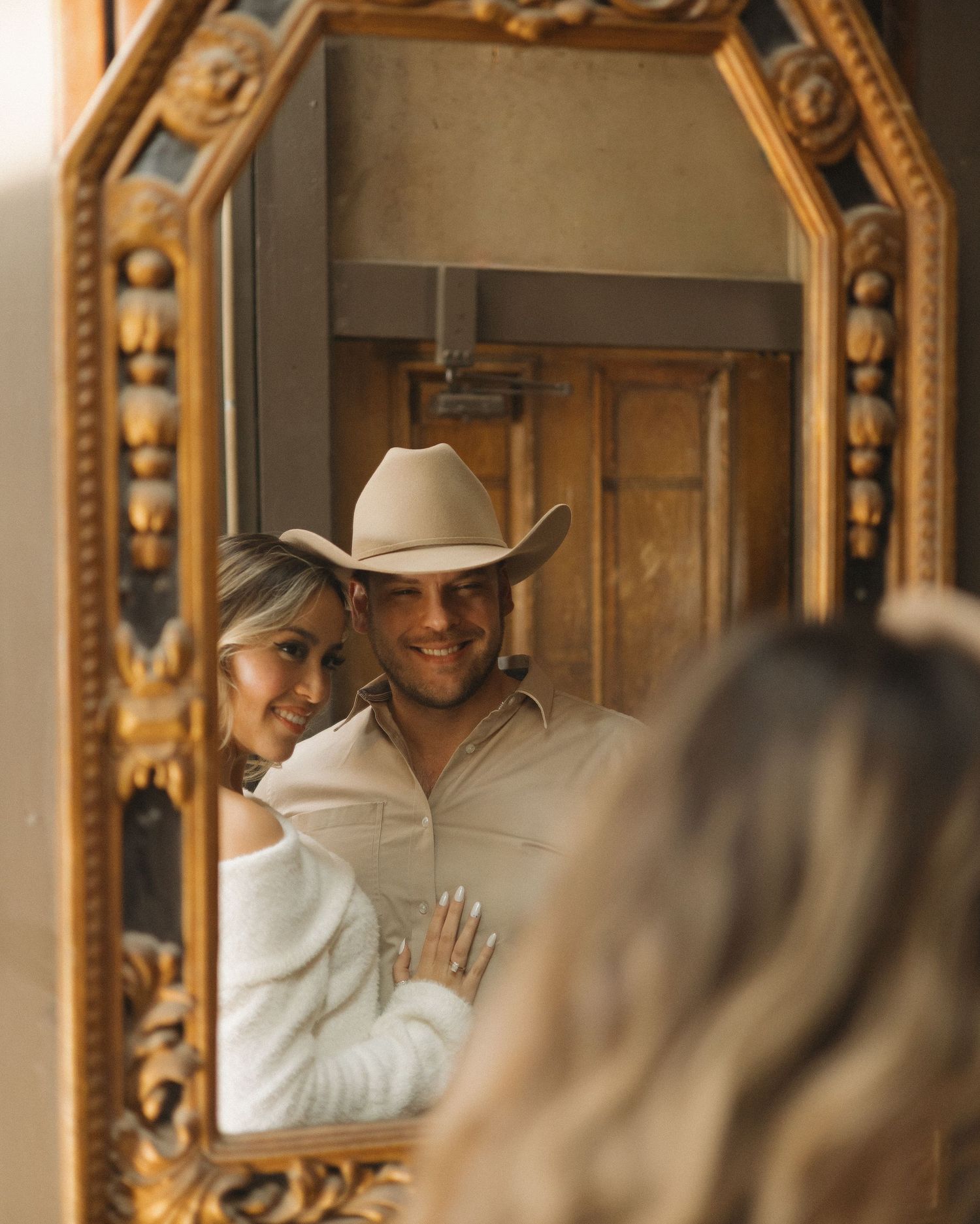Couple smiling in ornate gold mirror reflection, man wearing cowboy hat and woman in white off-shoulder top.