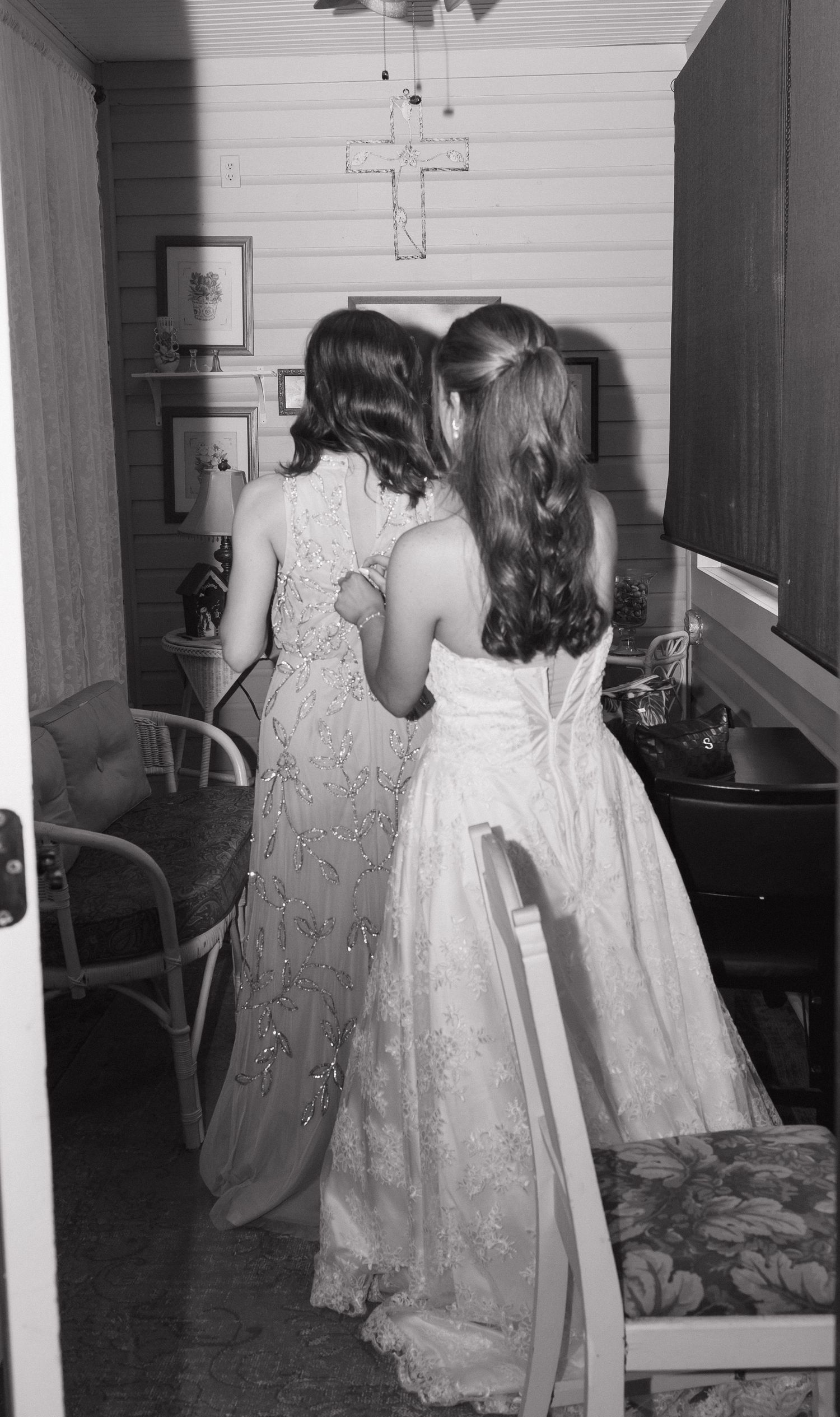 A vintage black and white photograph shows two people in long lace dresses standing in a room with a cross on the wall.