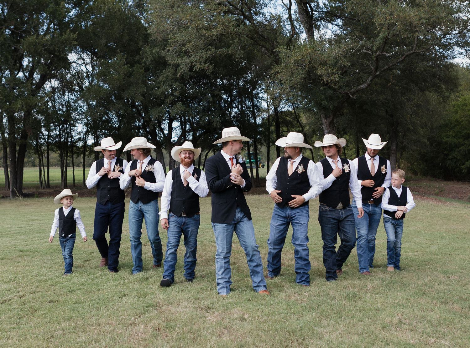 Wedding party in western wear with cowboy hats pose playfully in outdoor setting.