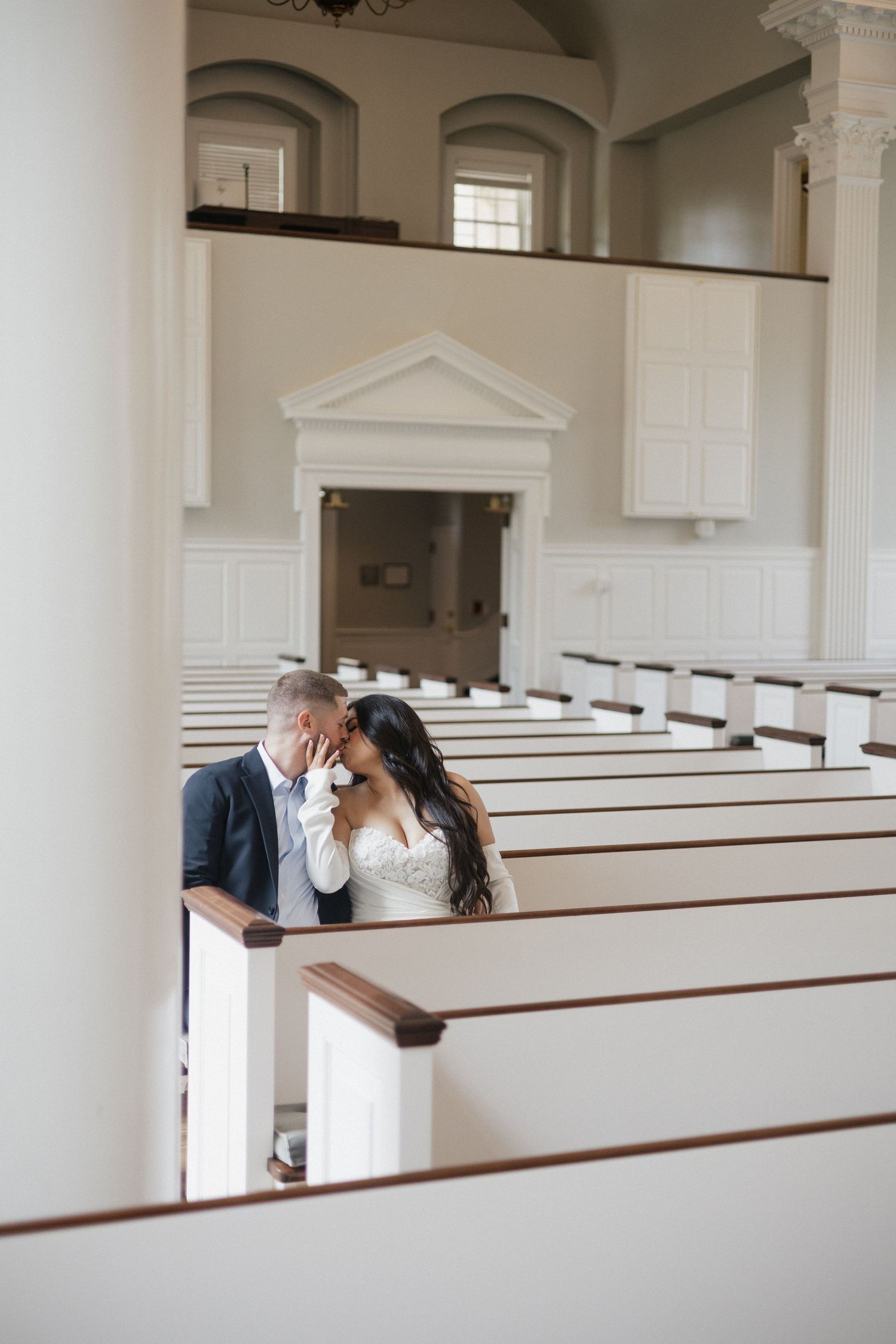 Bride and groom share a tender kiss between white church pews in an elegant chapel.
