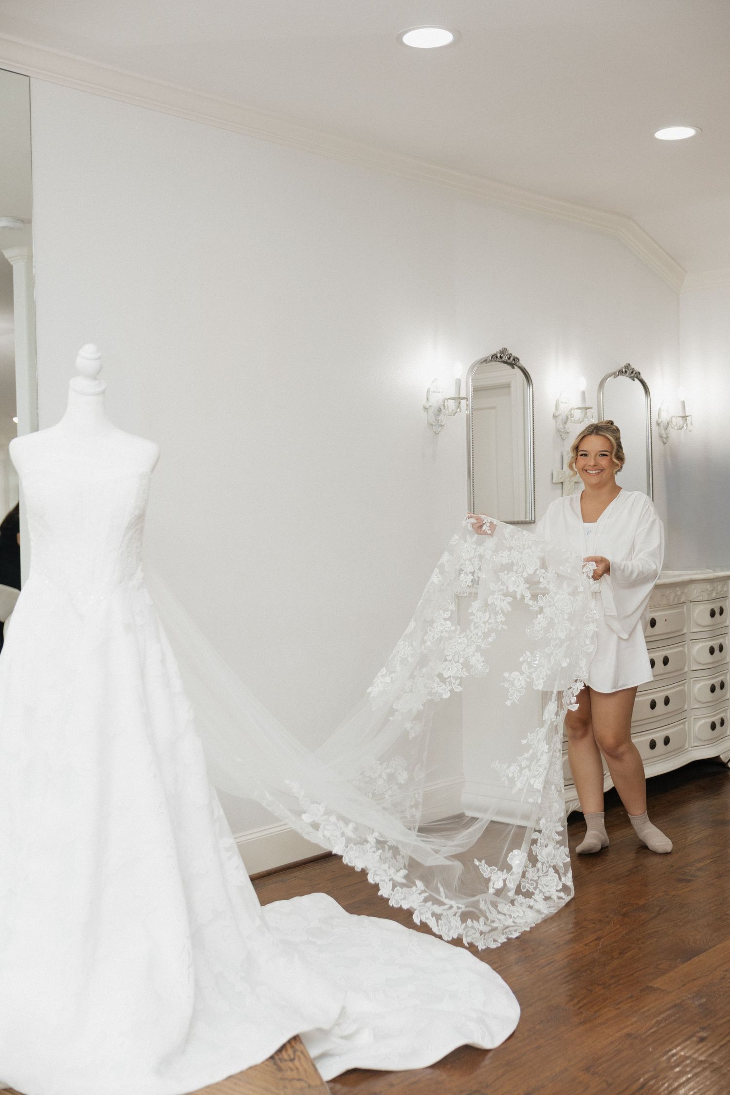 Bride in white robe holding lace veil next to wedding dress on mannequin in elegant bridal suite.