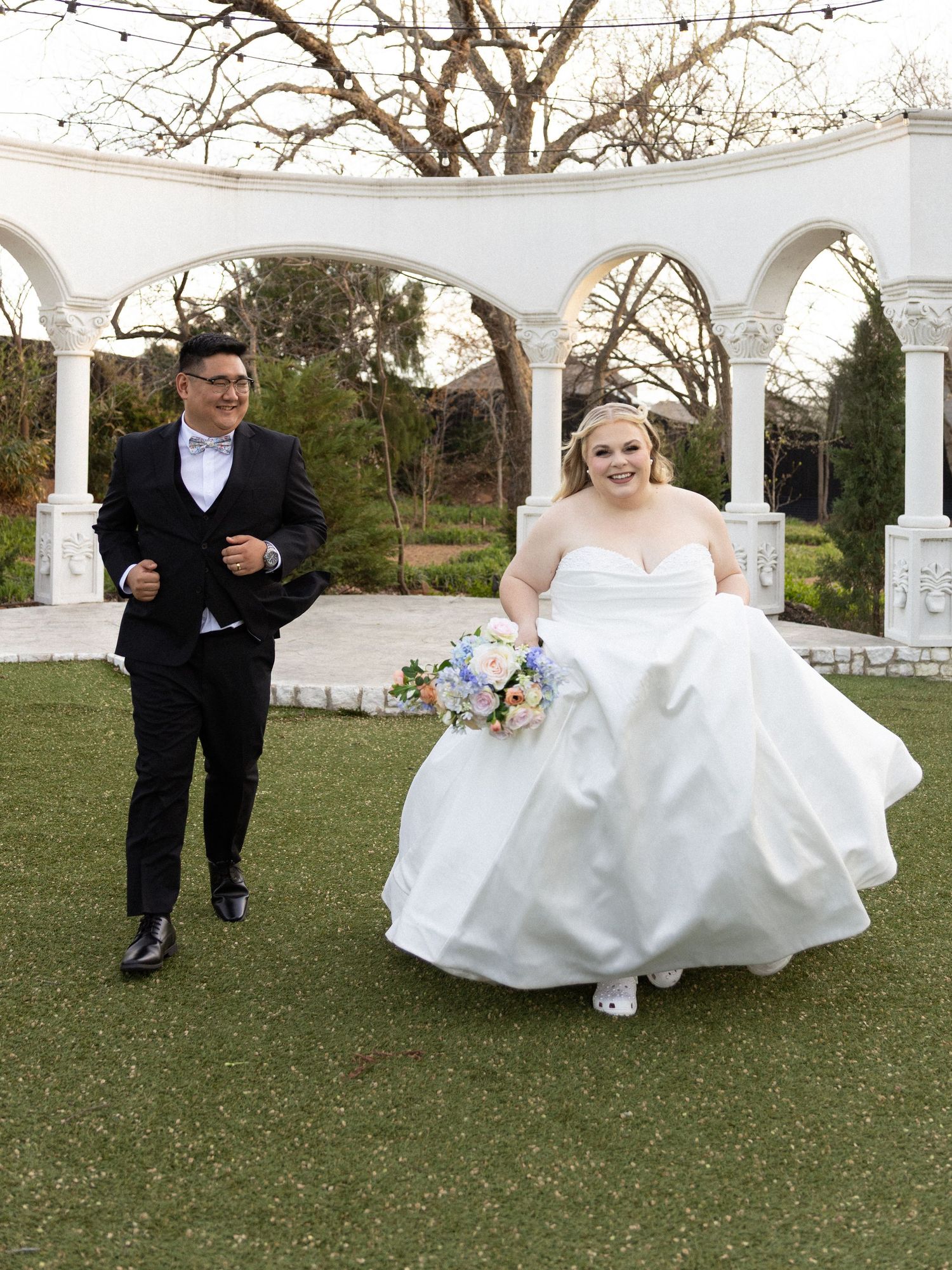 Bride in white gown holding bouquet walks with groom in black tux near elegant white arched pergola outdoors.