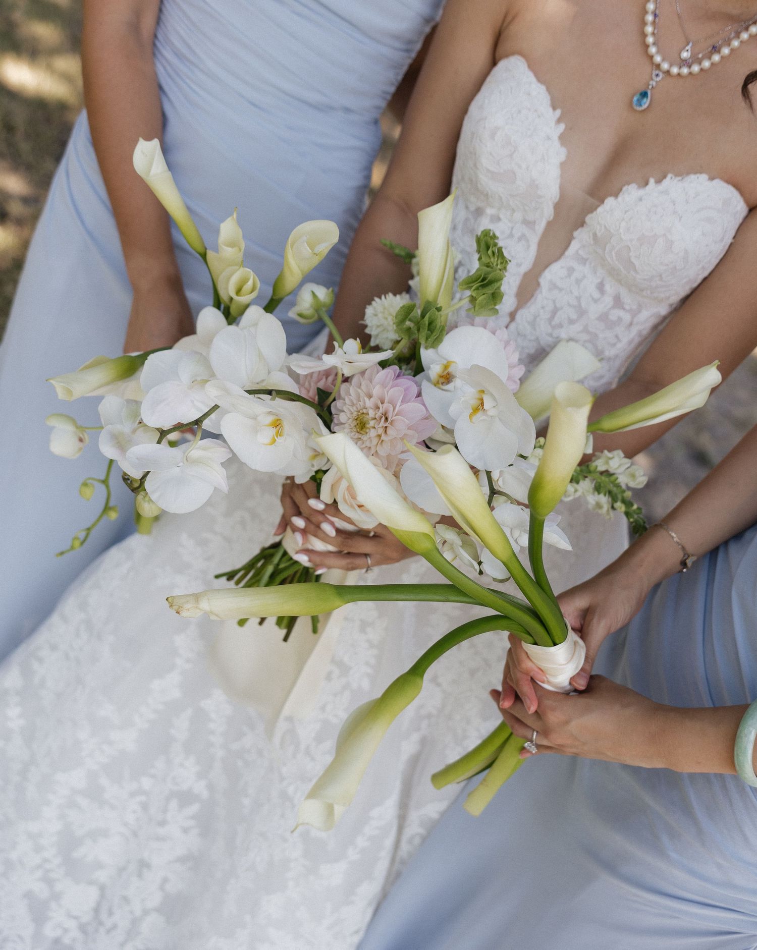 Bride in lace gown holds white calla lily and orchid bouquet beside bridesmaid in light blue dress.