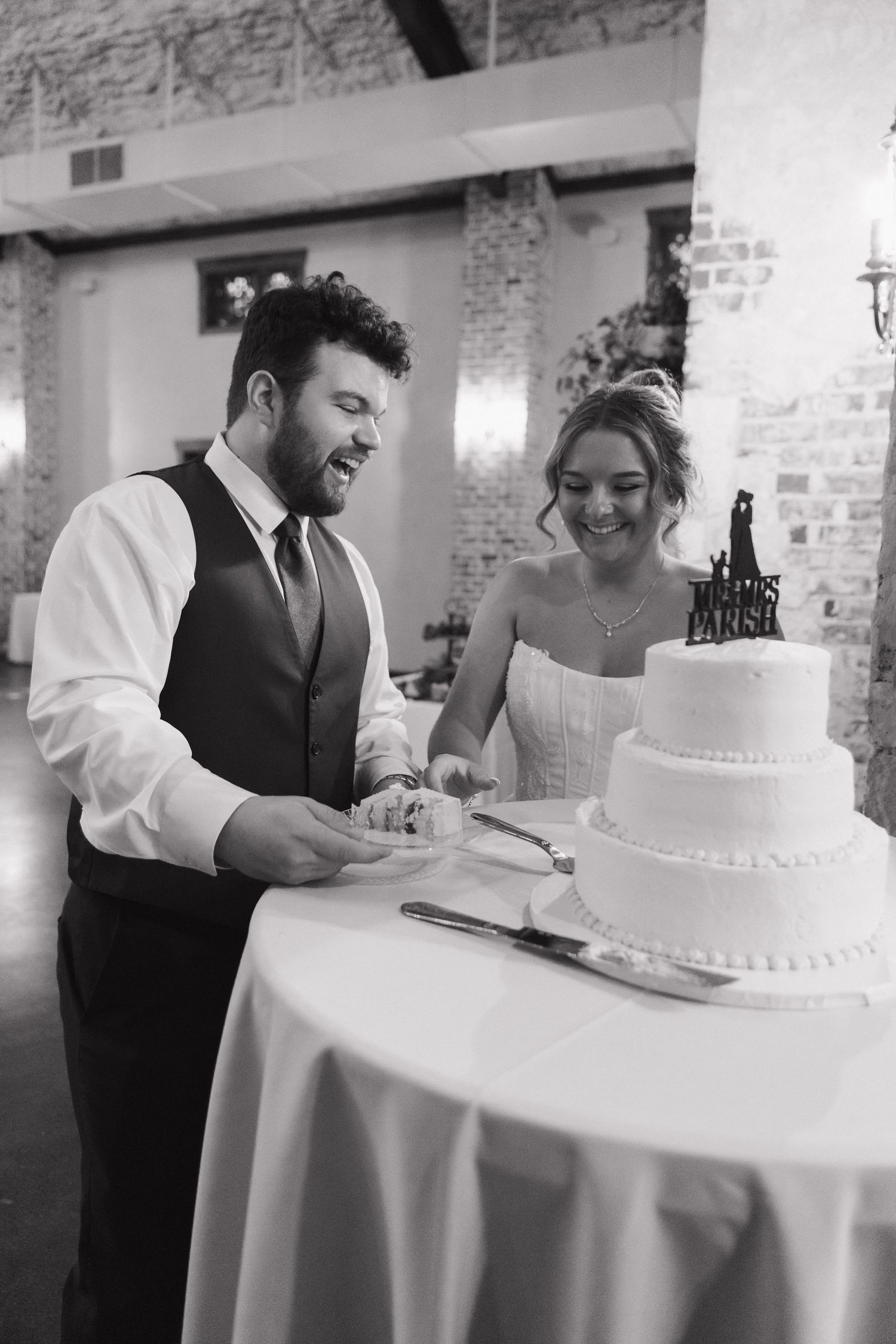 Bride and groom laughing while cutting their white three-tier wedding cake at reception.