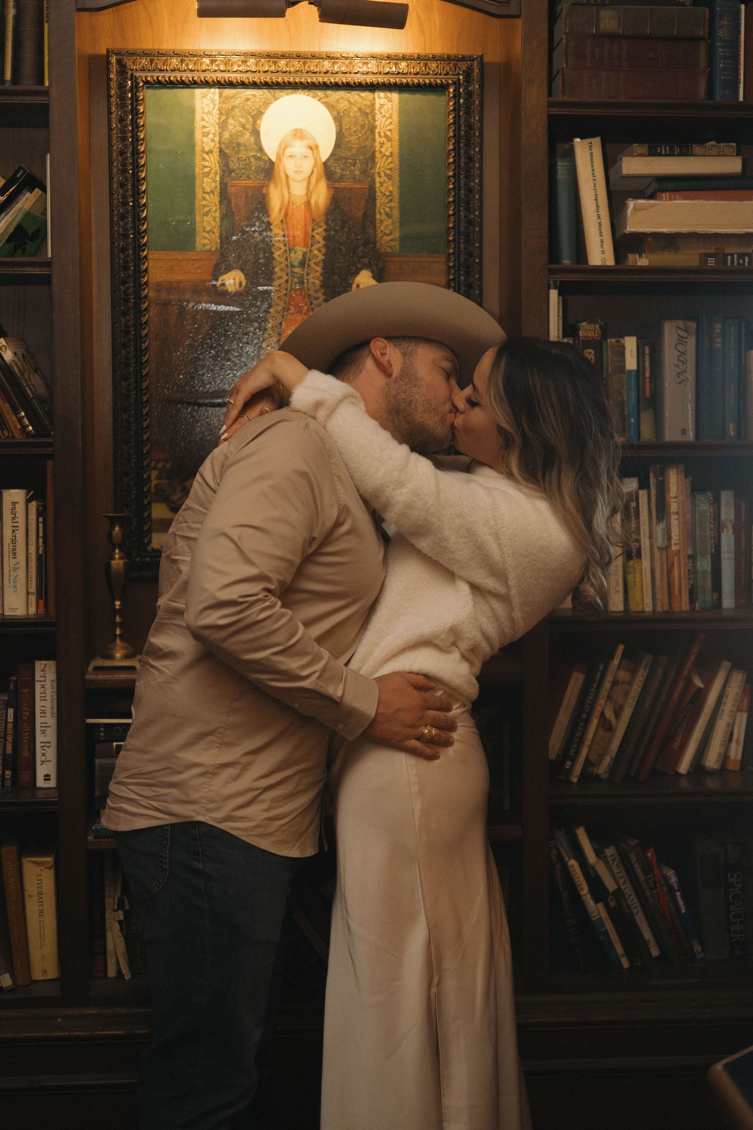 Couple sharing a romantic kiss in a cozy library with bookshelves and a glowing portrait painting behind them.