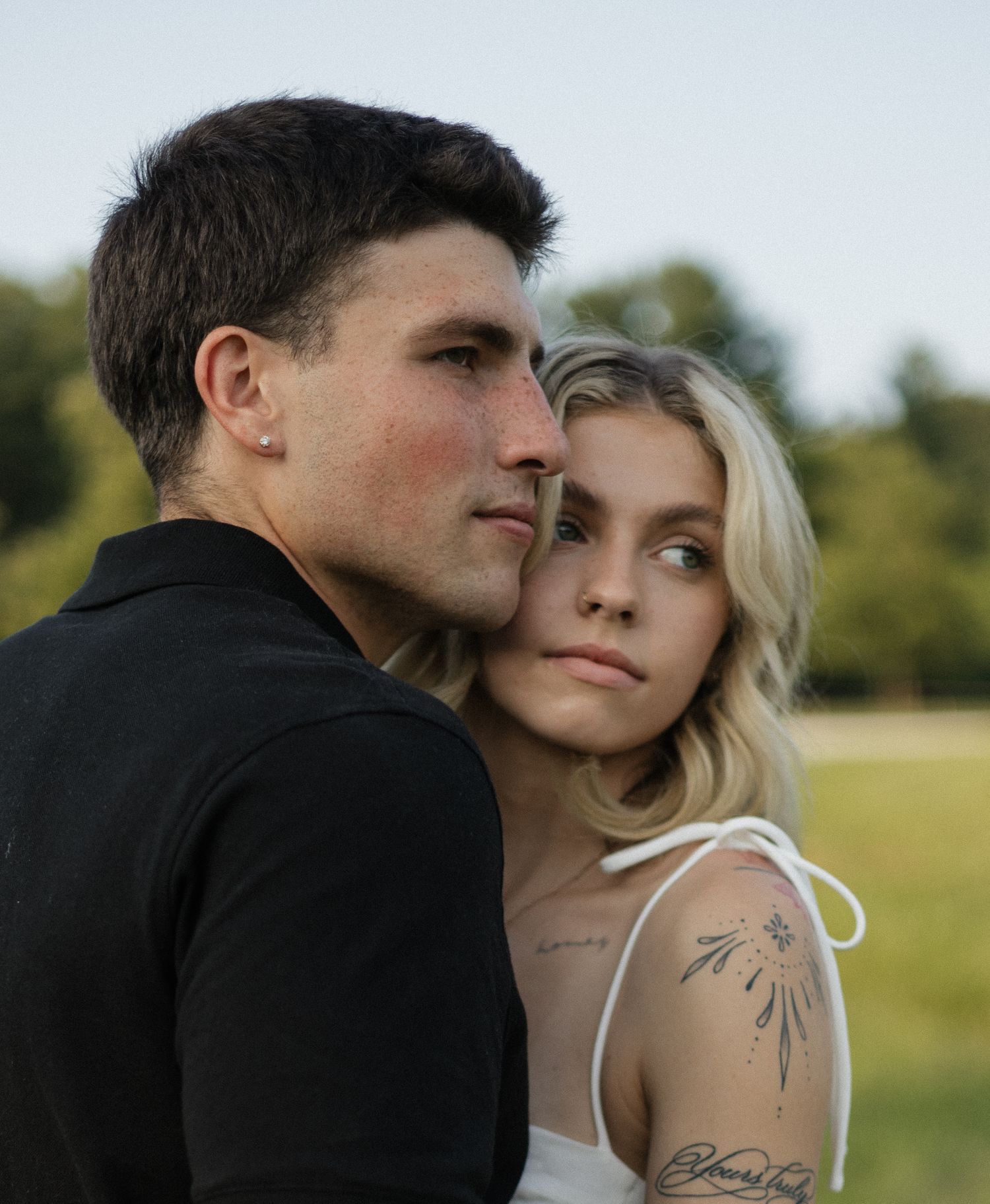 A young couple in an outdoor setting, man in black shirt and blonde woman with tattoos sharing an intimate moment.