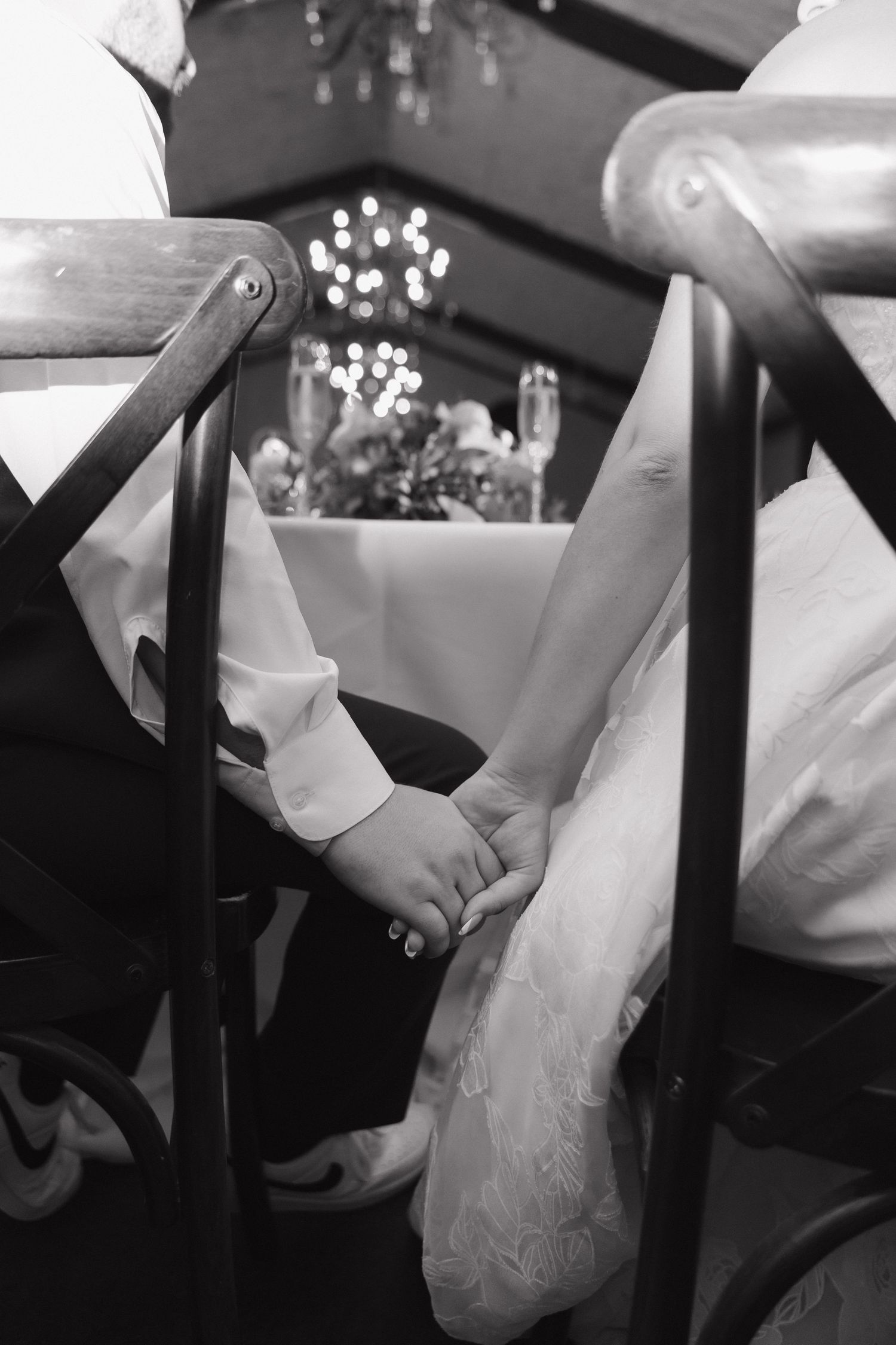 Black and white photo of bride and groom holding hands during wedding ceremony with chandelier in background.