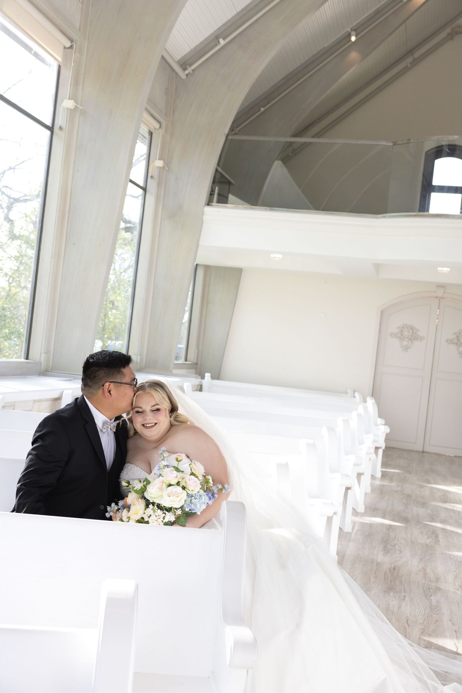 Bride and groom share a romantic kiss among white chapel pews, bride holding a pastel bouquet.
