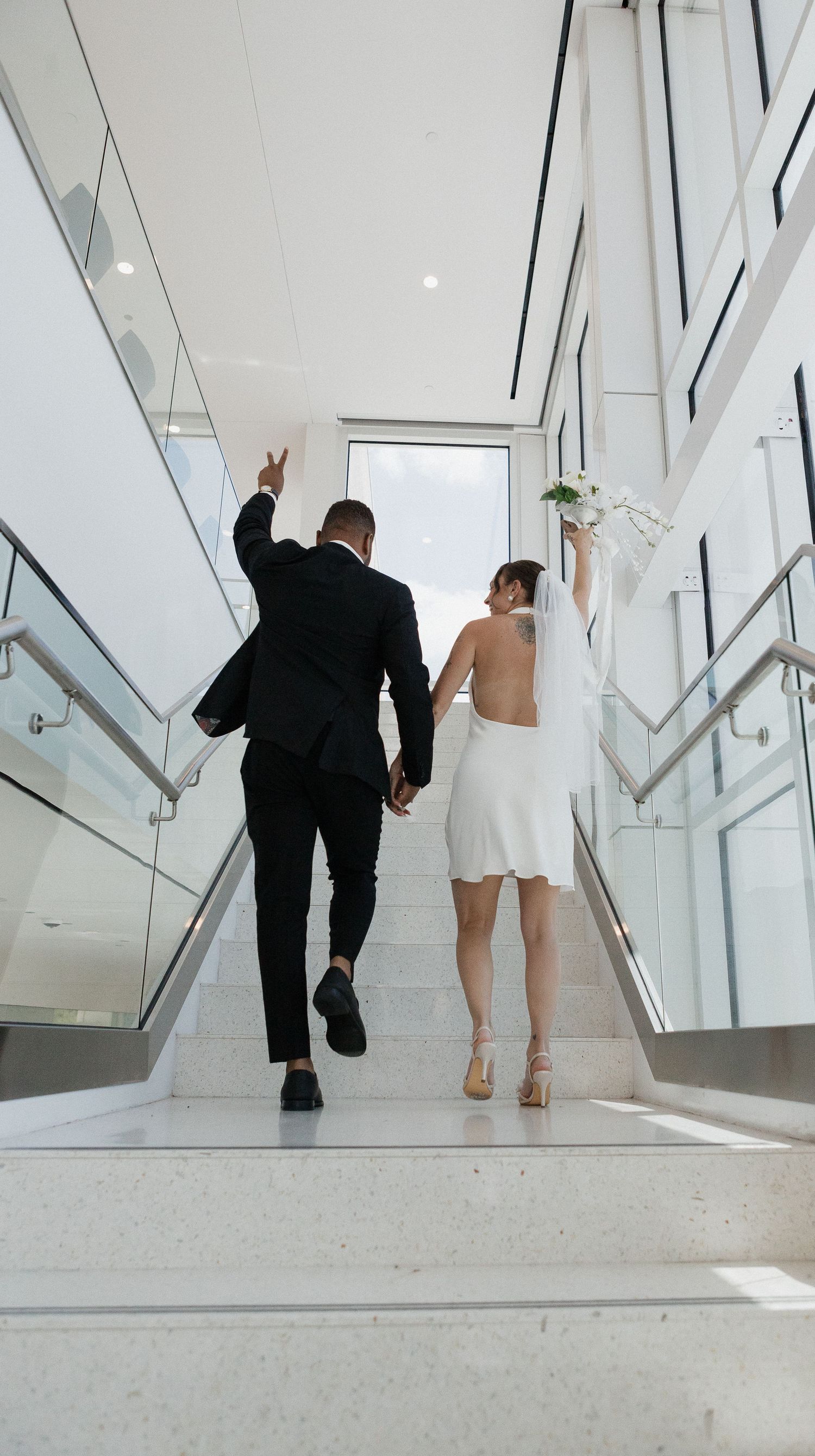 A couple in formal attire walks through a modern white hallway with angular geometric walls and natural lighting.
