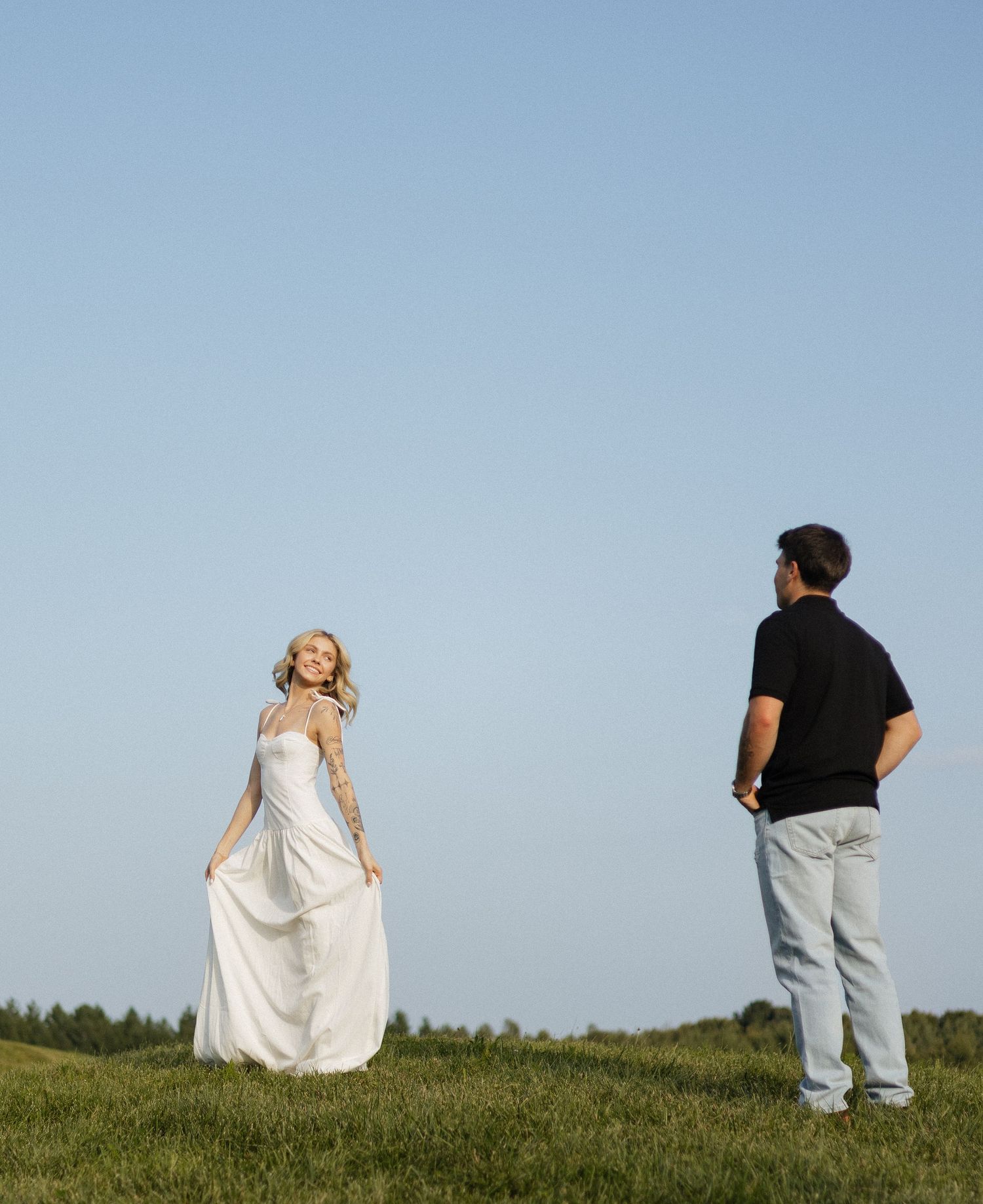 Bride in white gown and groom in black shirt stand on green hill under clear blue sky.