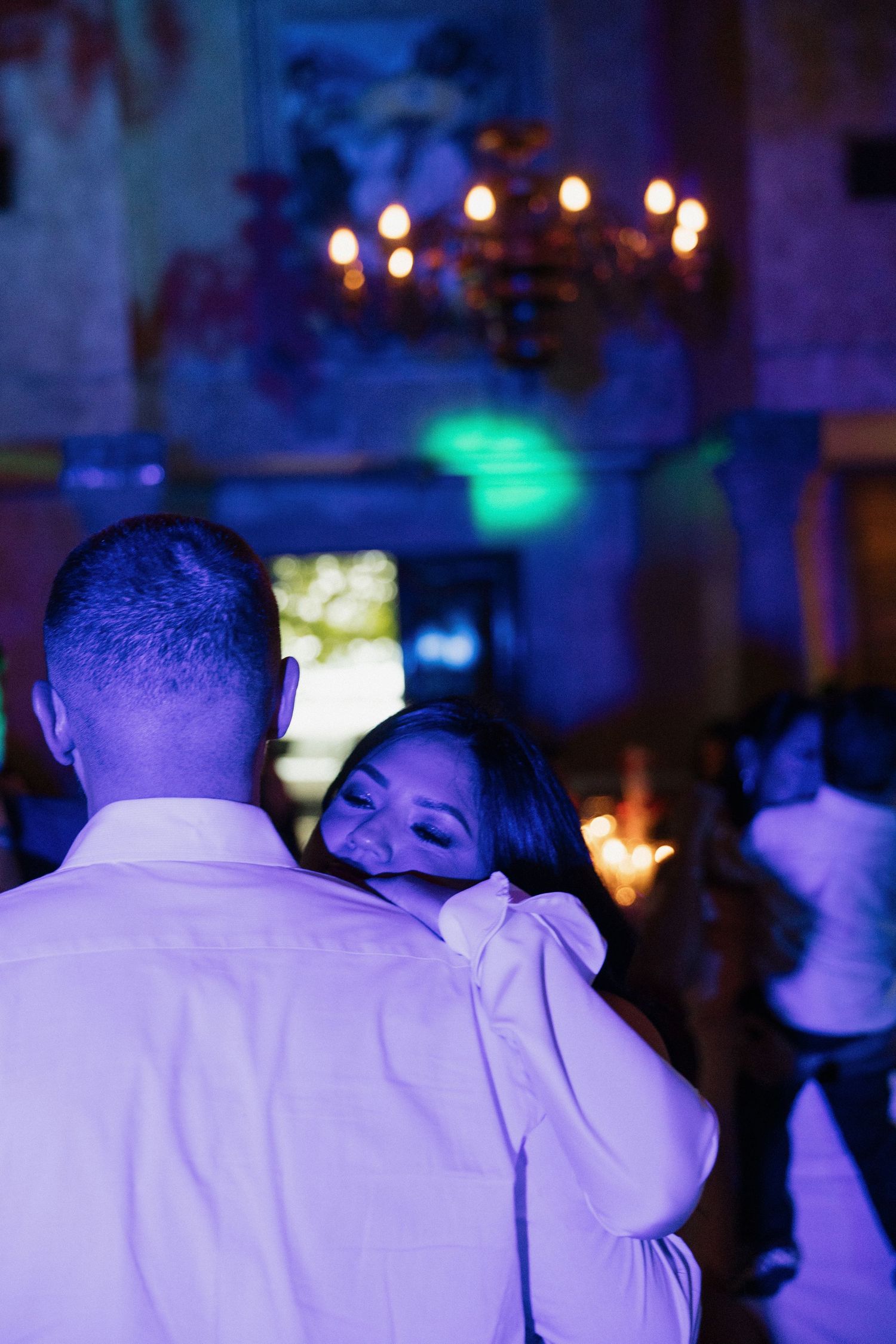 Bride resting head on groom's shoulder during romantic first dance under chandelier with blue reception lighting.