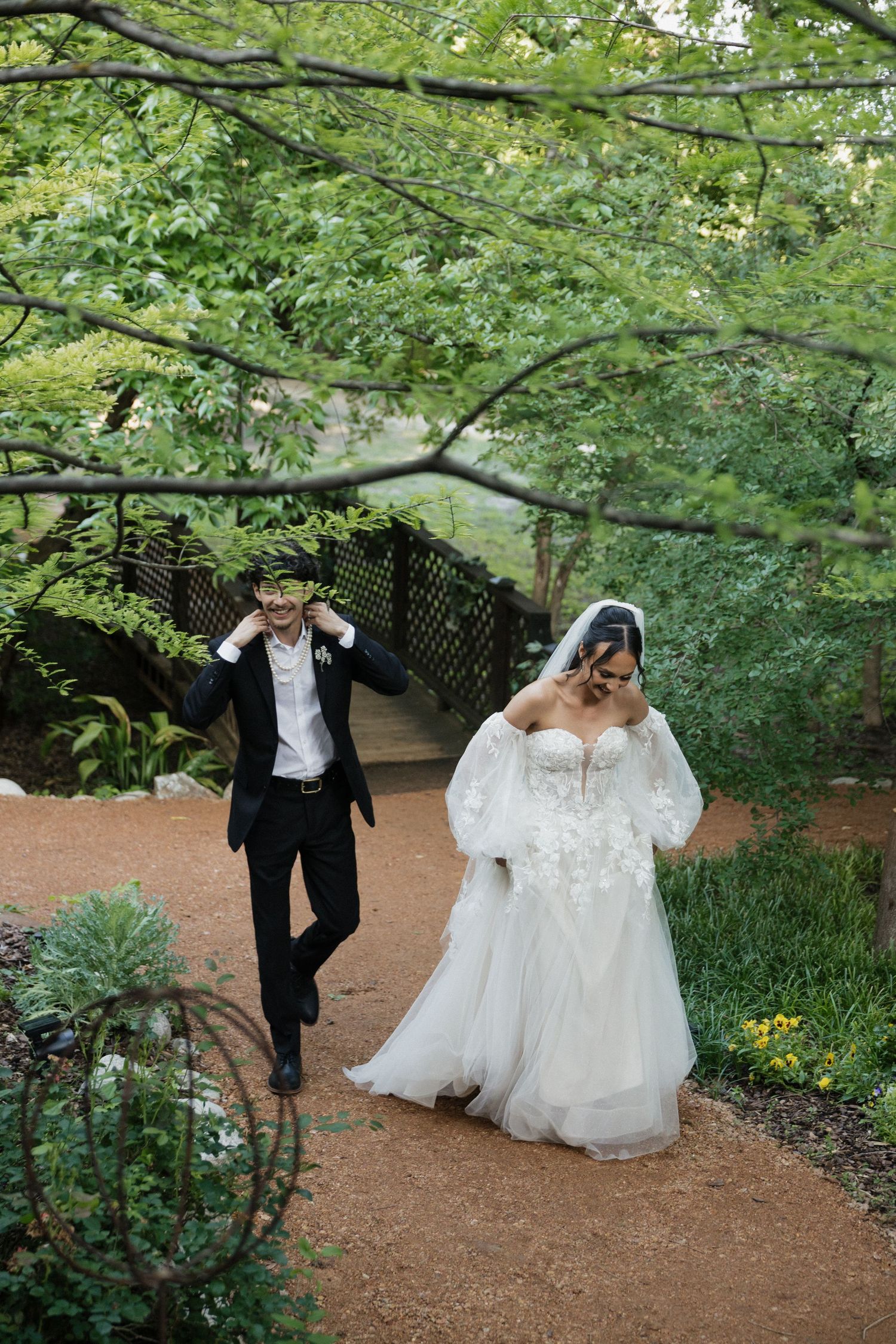 Bride in white lace gown and groom in black tuxedo laughing together on a garden path surrounded by lush greenery.