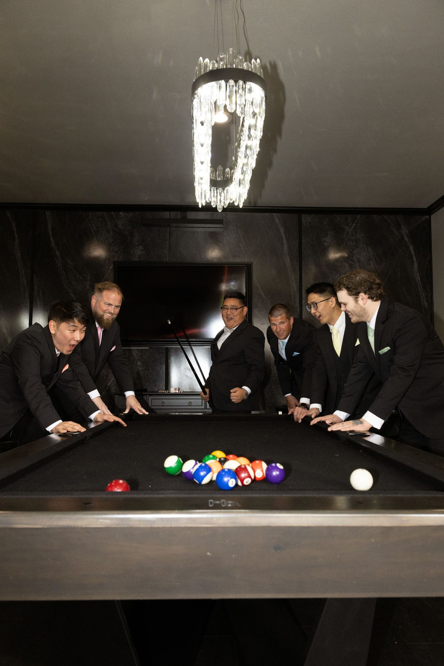 Seven groomsmen in black suits gather around a pool table with colorful billiard balls under a crystal chandelier.