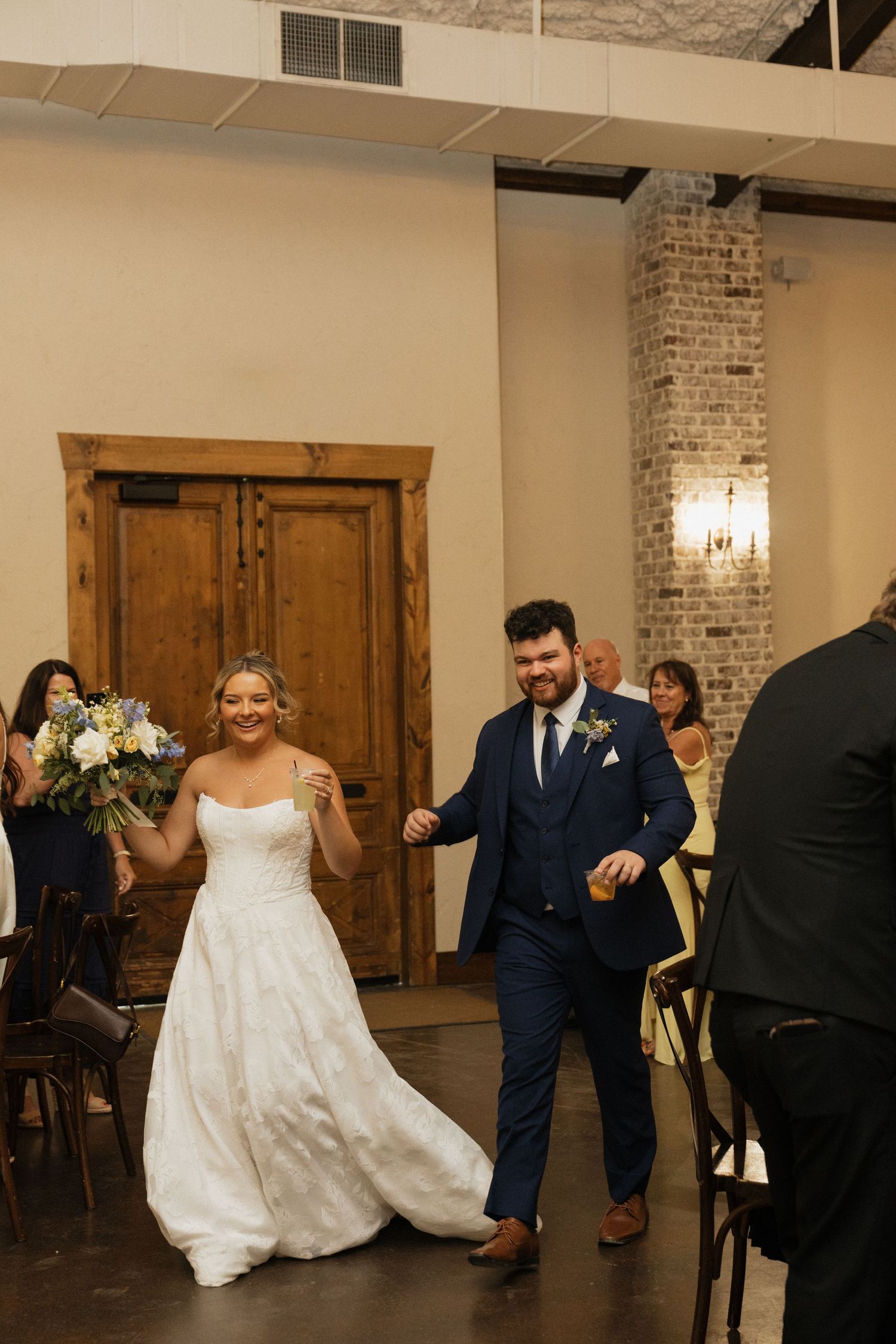 Bride and groom walk joyfully into wedding reception hall, bride holding blue and white bouquet in elegant white gown.