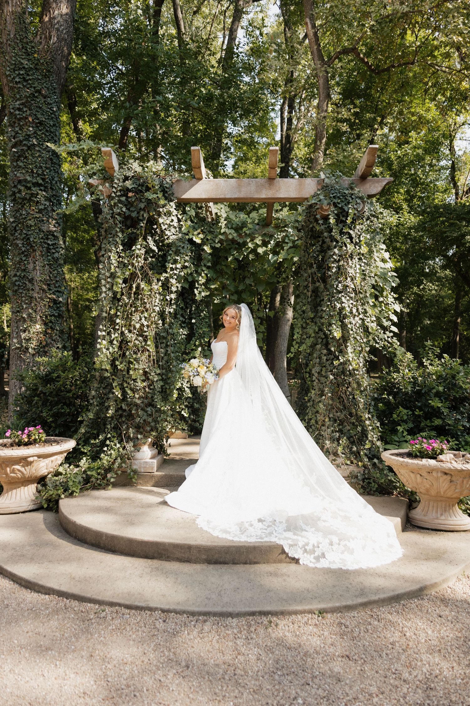 Bride in flowing white gown and cathedral veil stands beneath ivy-covered arch in lush garden setting.