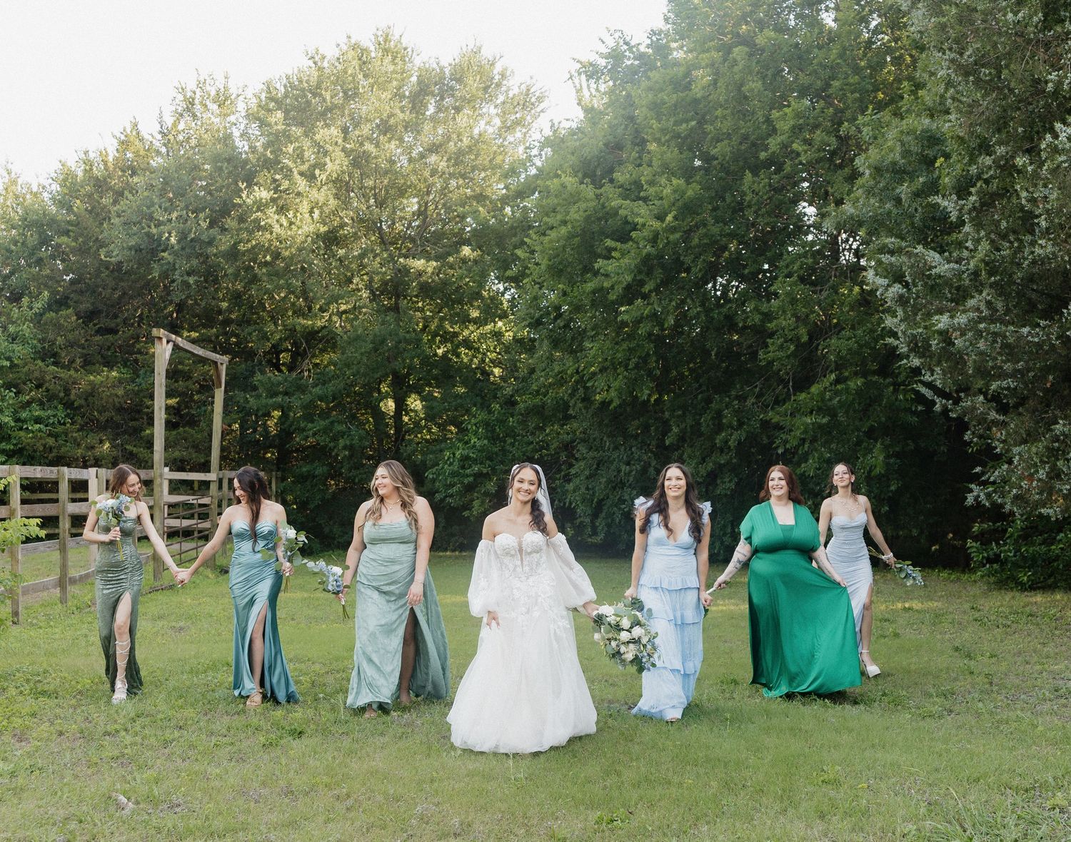Bride in white gown walks with six bridesmaids in green dresses on a lush outdoor lawn at a wedding.