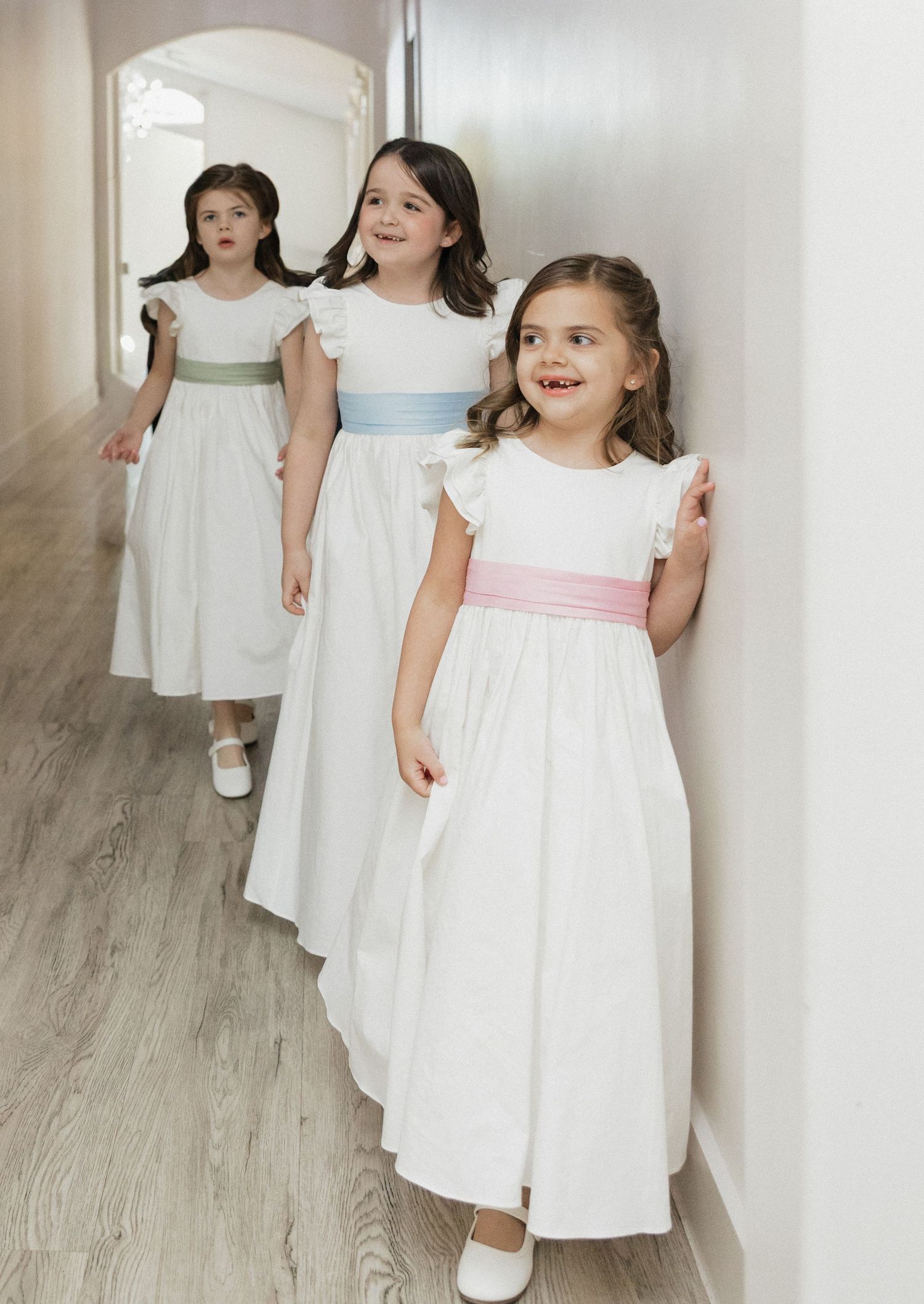 Three girls in white dresses with colorful sashes in pink, blue, and green walk down a bright hallway.