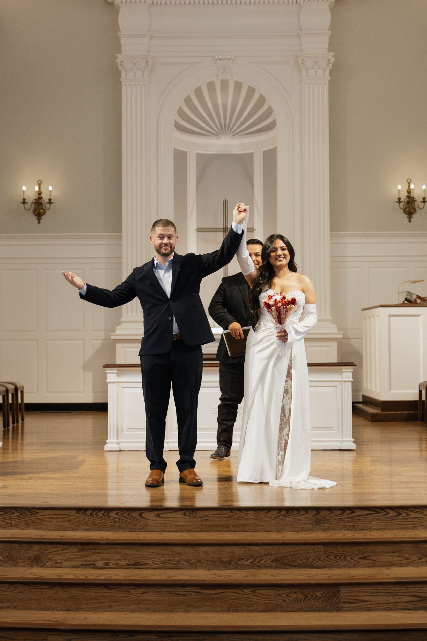 Joyful newlyweds celebrate with officiant at church altar after wedding ceremony.