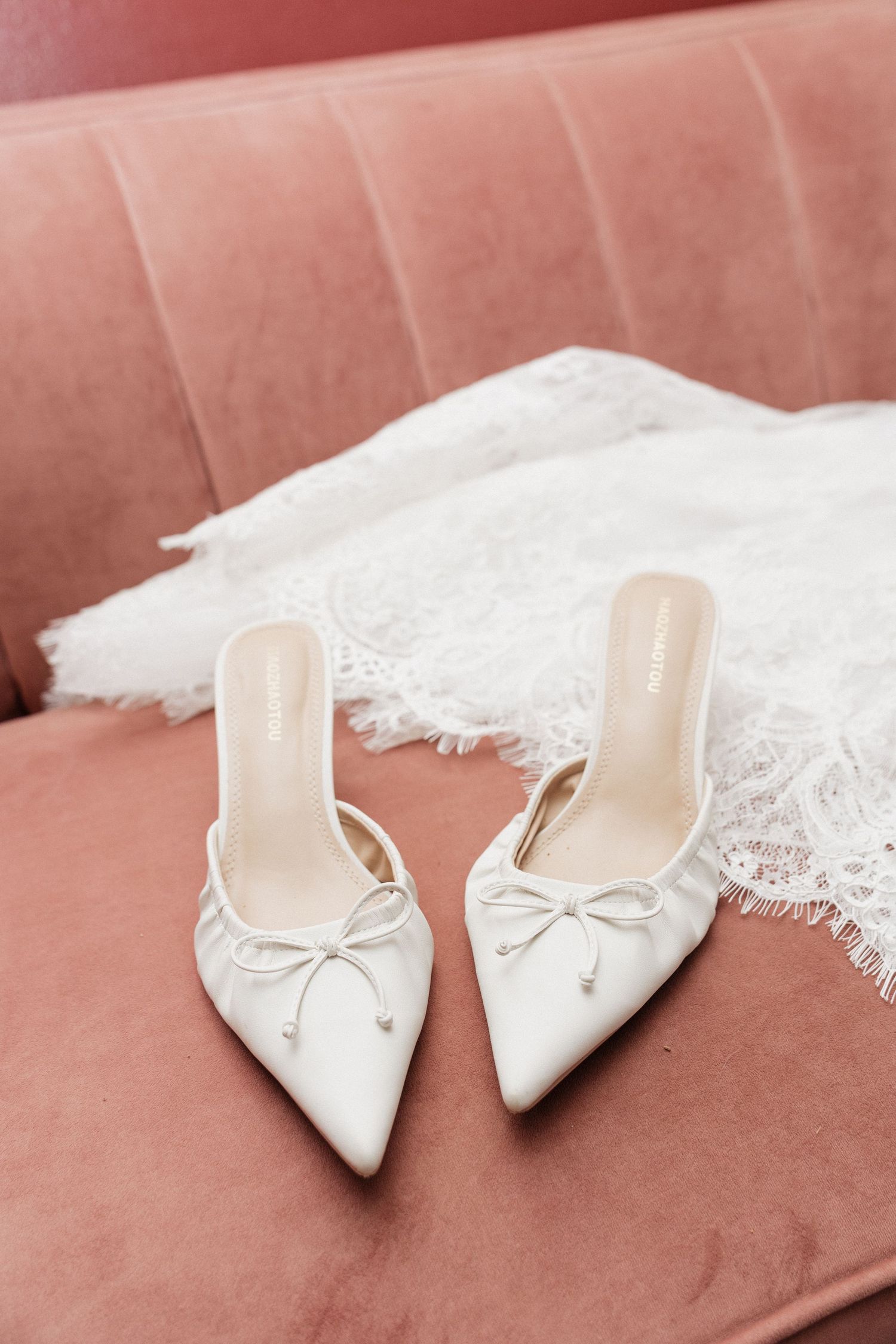 White satin bridal mules with bow detail on pink velvet sofa beside lace wedding dress.