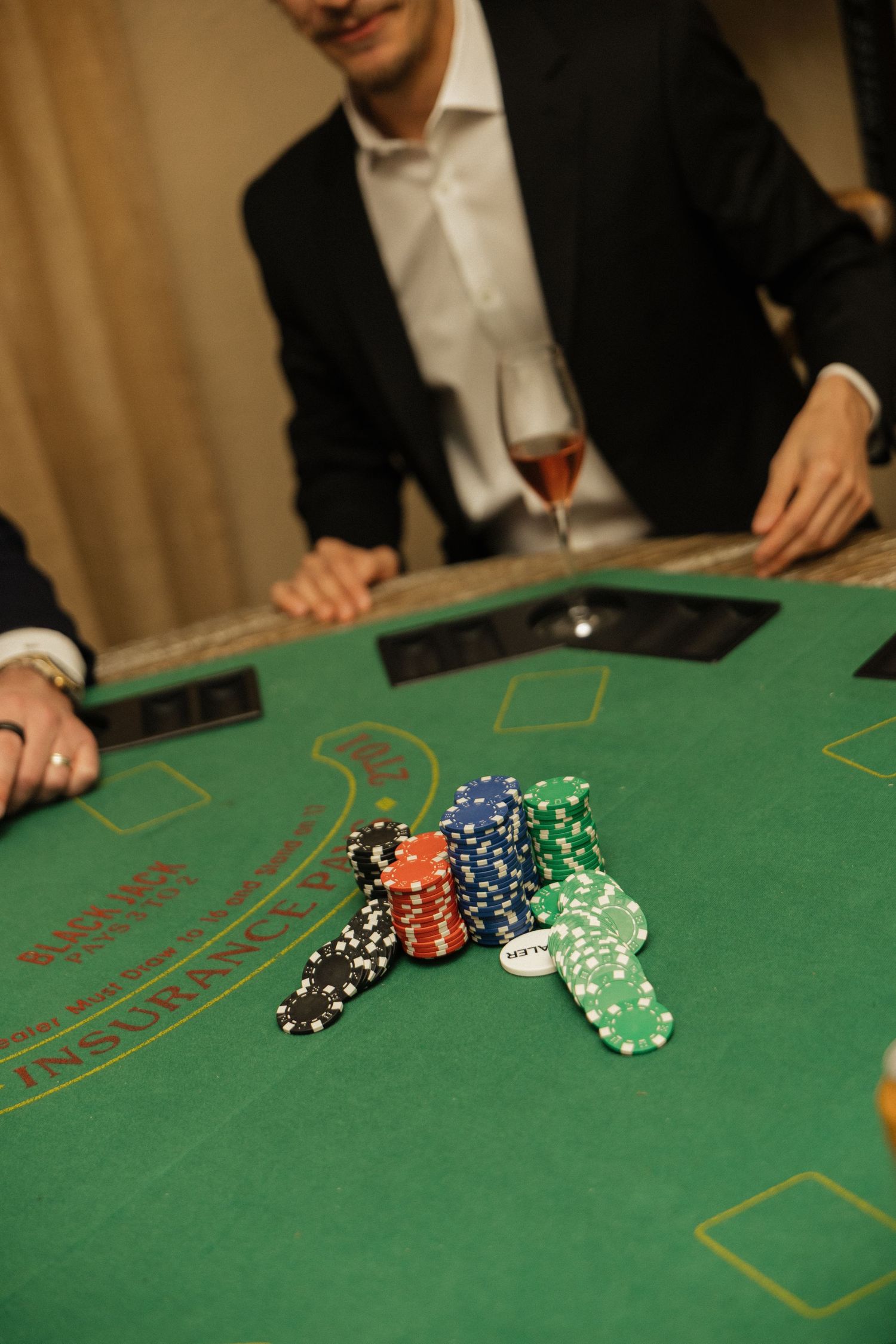 Colorful casino chips stacked on a green blackjack table with players in formal attire in the background.
