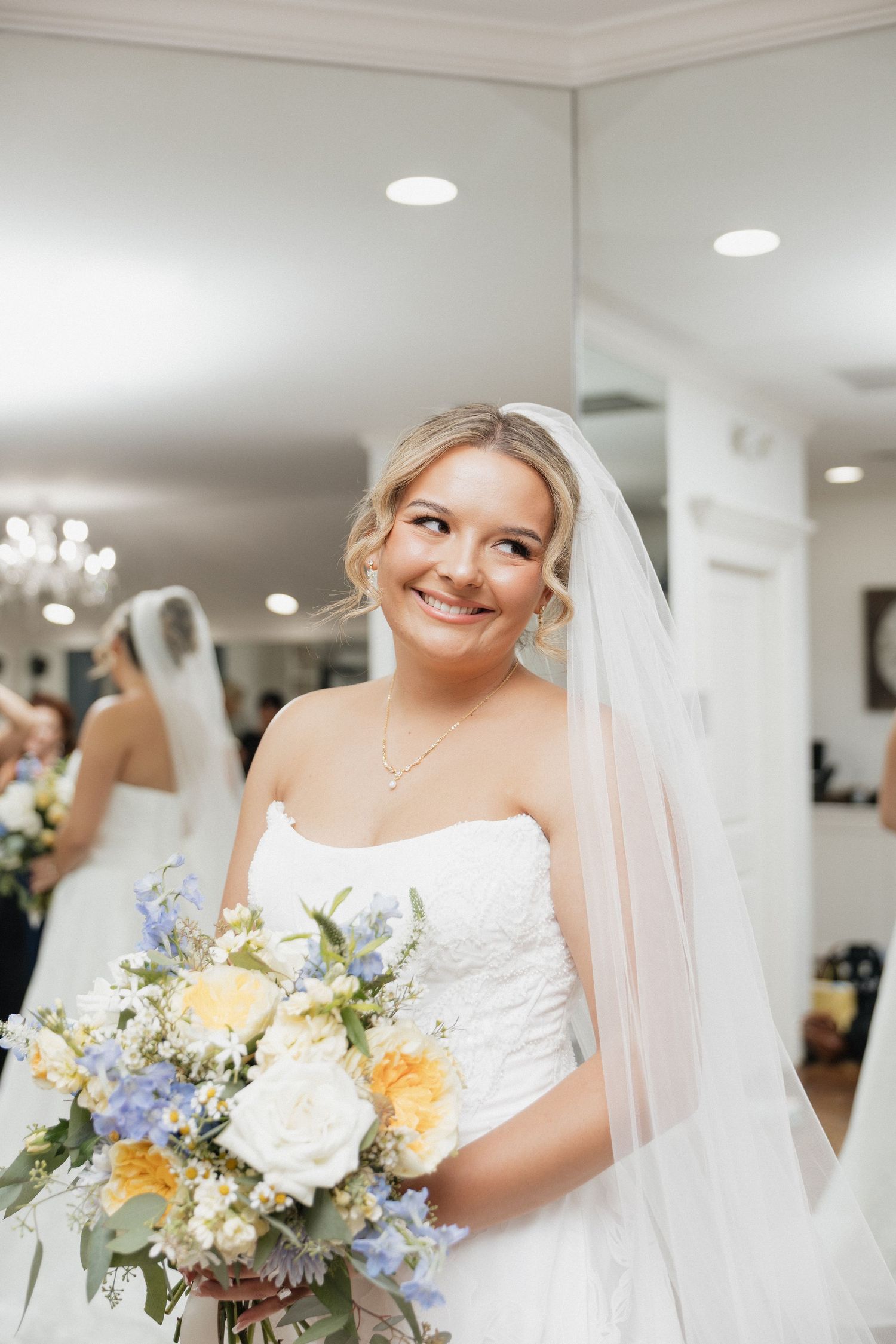 Smiling bride in strapless lace gown holds yellow and blue bouquet while wearing veil in bright bridal suite.