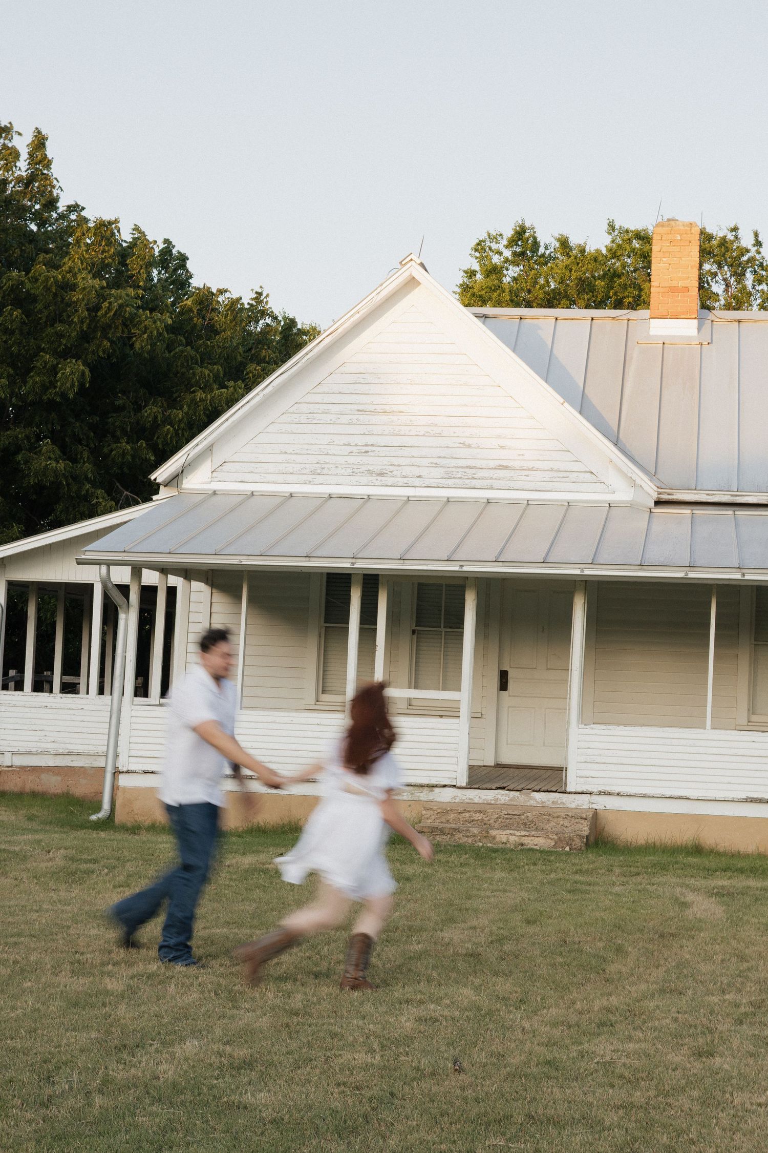 Couple holding hands and running in front of a white farmhouse with a metal roof and covered porch.