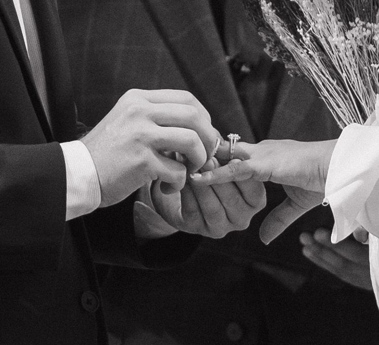 Black and white photo of a groom placing a diamond engagement ring on a bride's finger during a wedding ceremony.