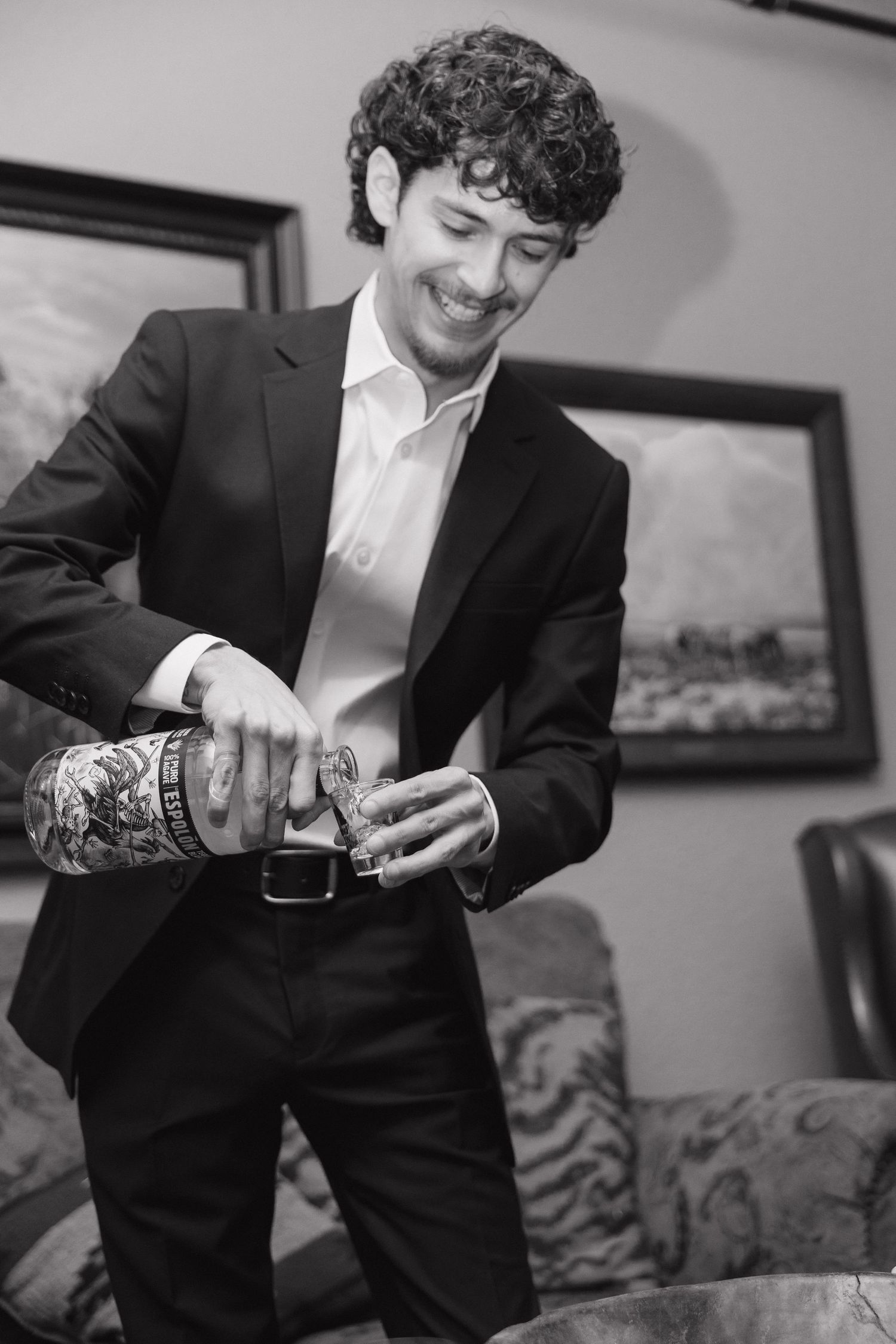 Smiling groom in black suit pouring a drink, black and white wedding photo with tattoos visible.