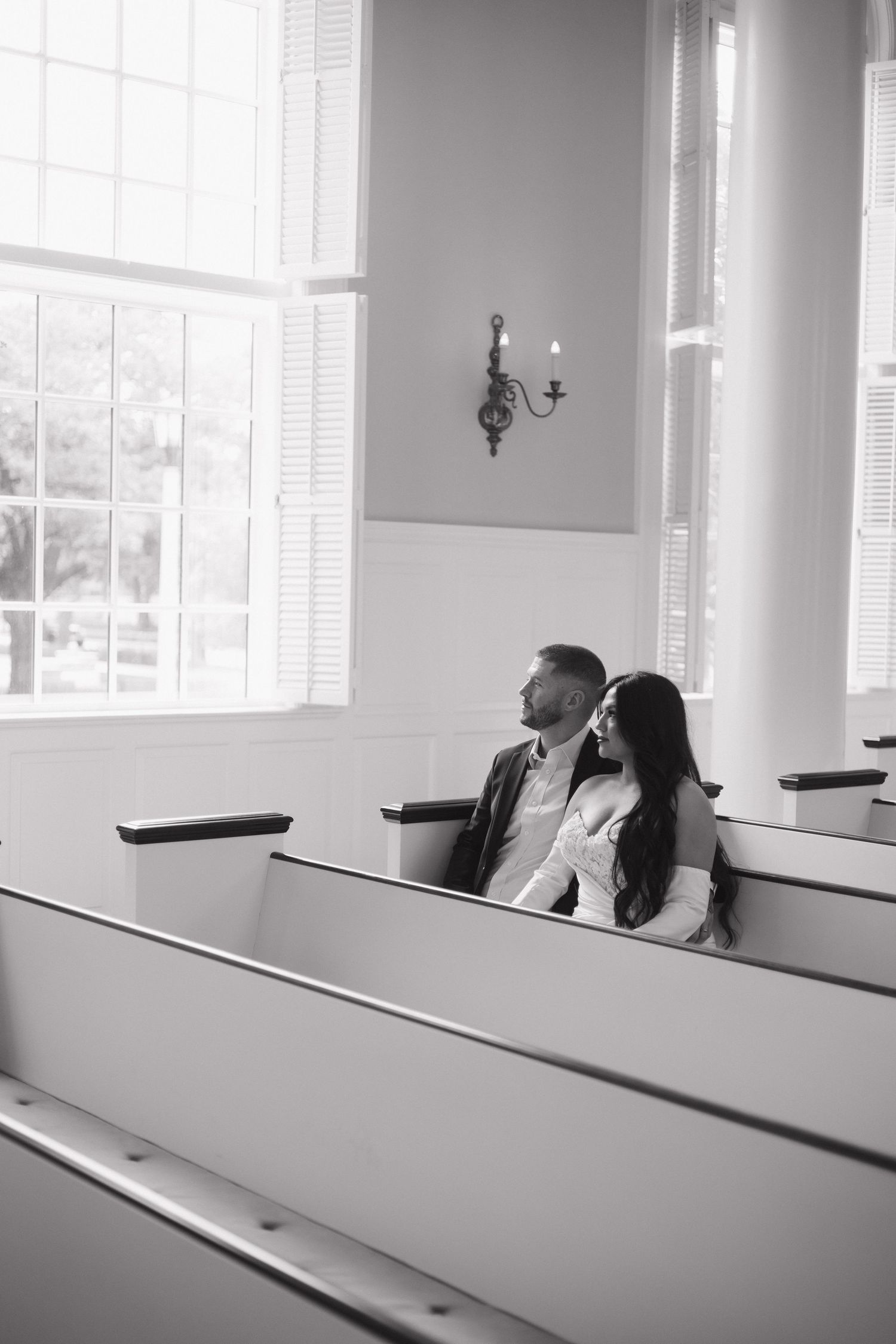 Couple sits together in church pew in black and white wedding photo.