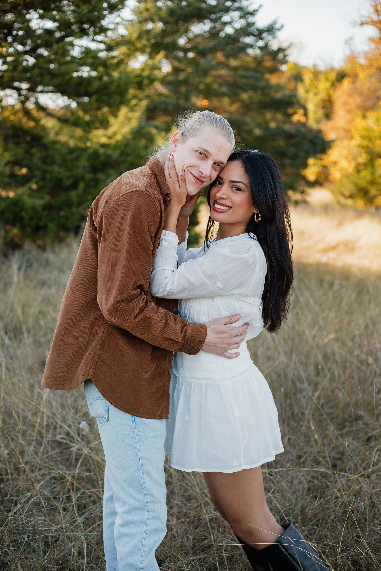 Intimate moment captured between couple in white dress and brown jacket against autumn scenery.