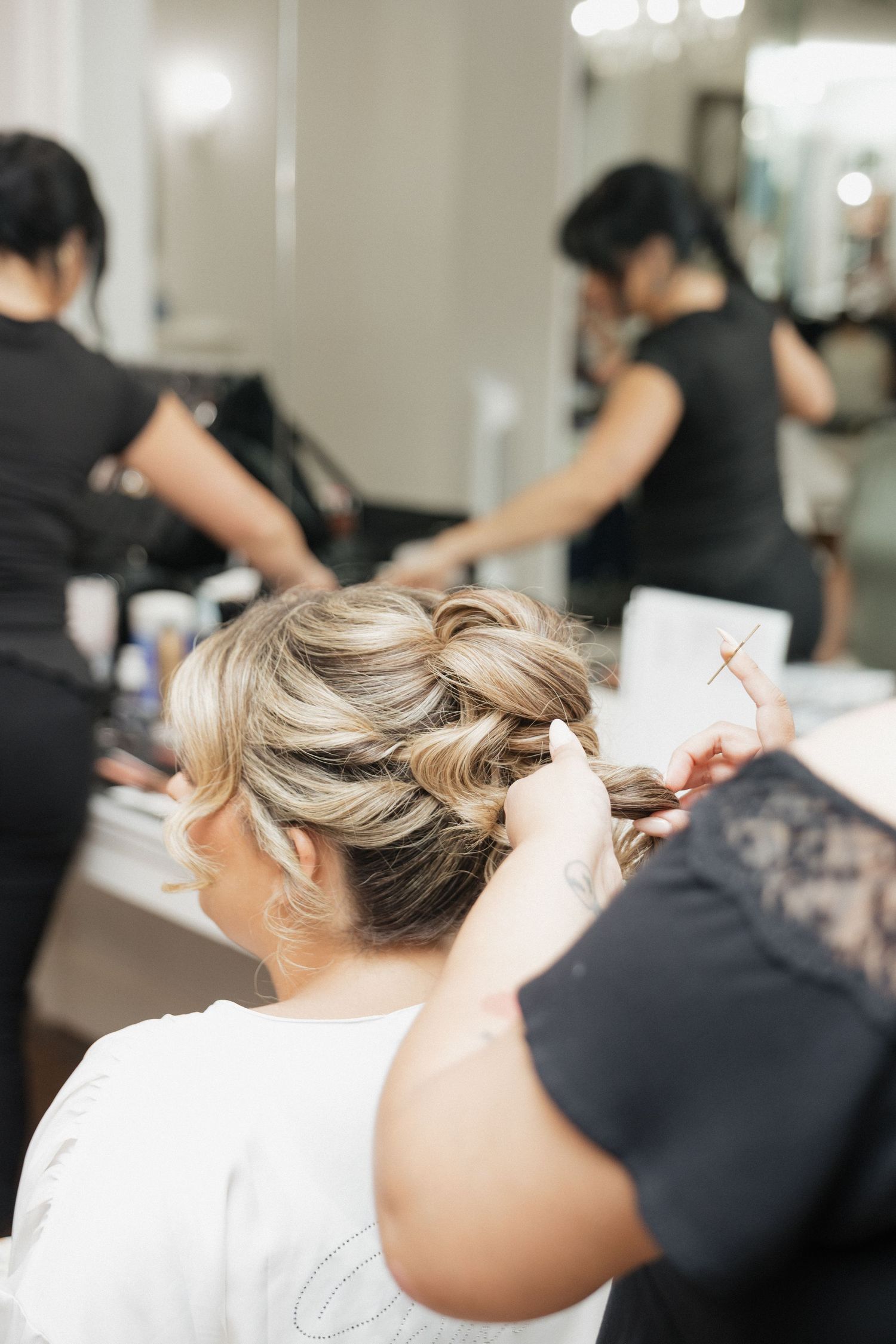 A hairstylist pins up a blonde updo hairstyle in a busy salon with mirrors and stylists in the background.