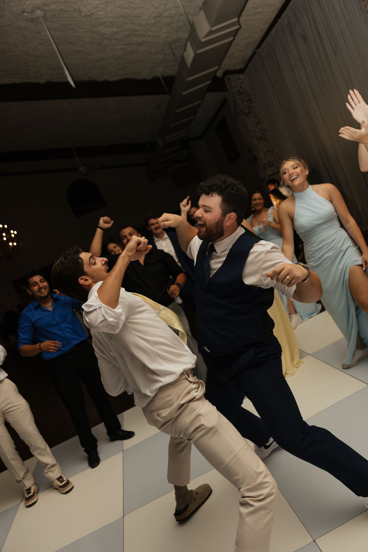 Groom in navy vest dances energetically with guests on white checkered floor at wedding reception.