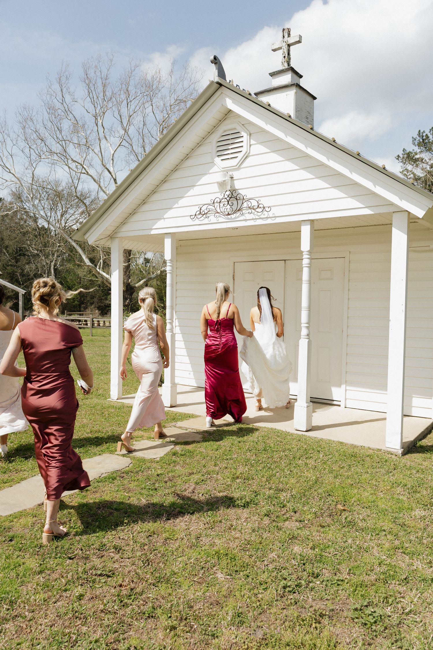 A group in formal dresses walking towards a small white chapel with a cross on top during a sunny day.