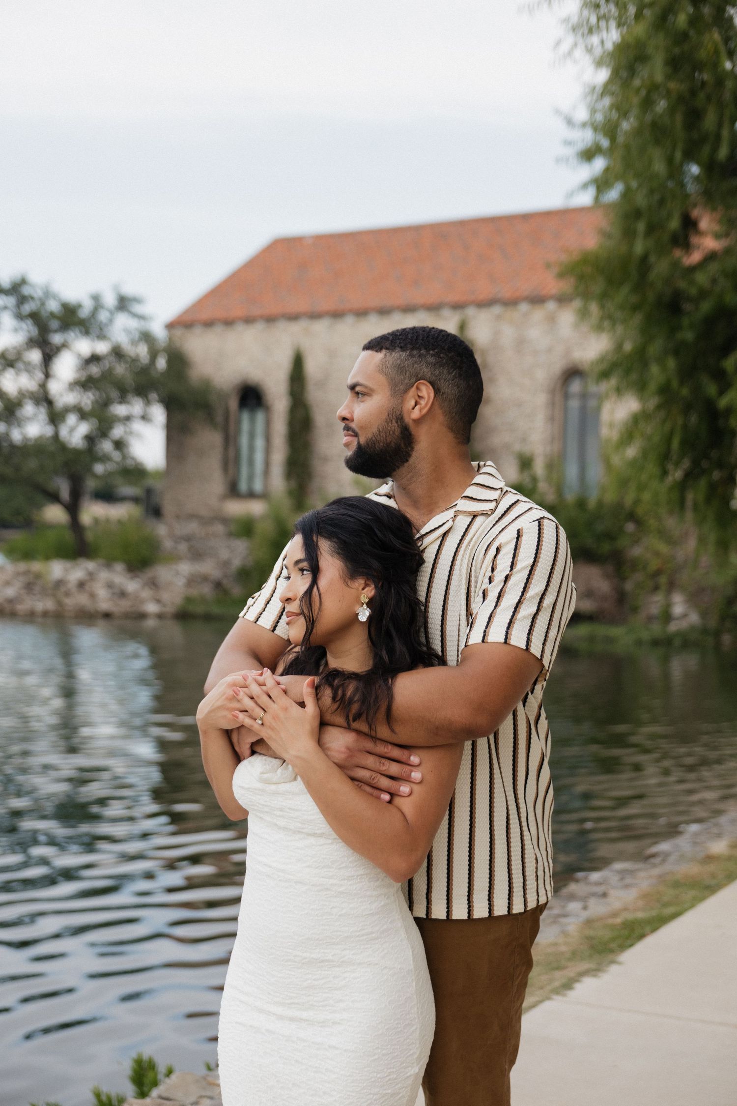 Couple embracing by a lake near a stone building, woman in white dress, man in striped shirt.