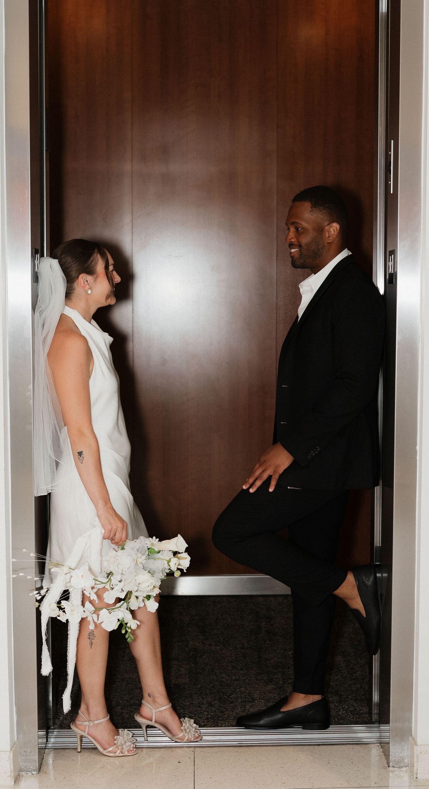 A couple shares a candid moment in a doorway, with the bride in a white dress holding a bouquet and groom in black suit.