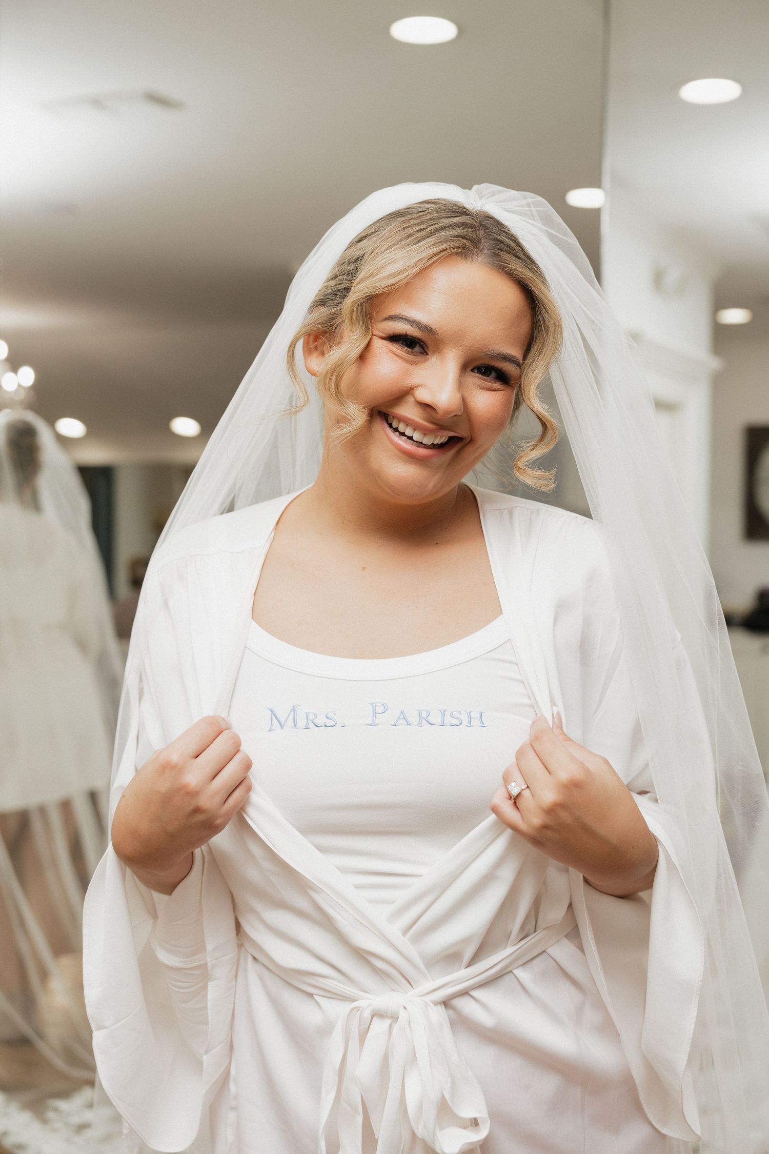 Smiling bride wearing a veil and white robe, showing off a Mrs. Parish tank top in a bridal boutique.