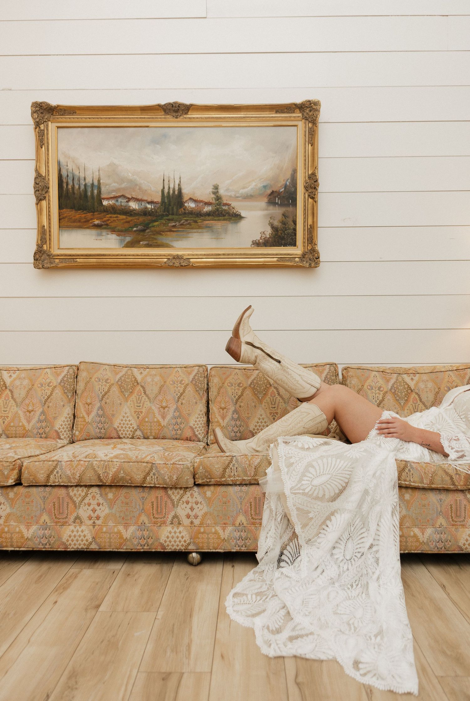 Bride in lace dress and tan cowboy boots lounging on vintage floral sofa beneath ornate gold-framed landscape painting.
