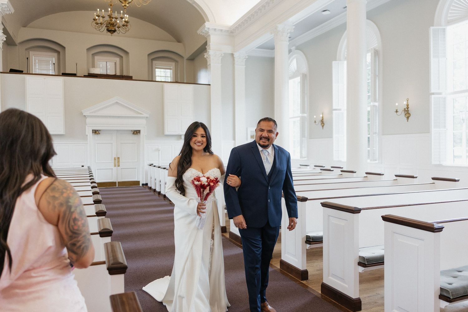 Bride in white gown walks down church aisle with groom in navy suit, holding red bouquet in elegant white chapel.