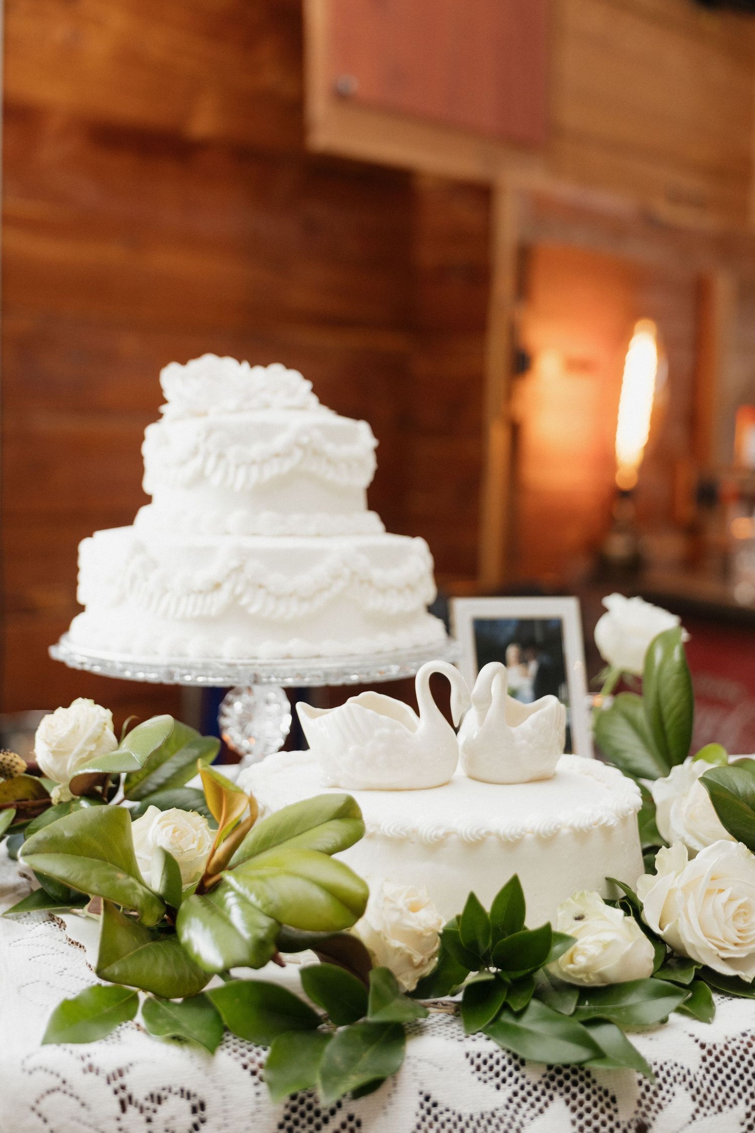 Elegant two-tier white wedding cake with swan toppers surrounded by white roses and magnolia leaves on a lace tablecloth.