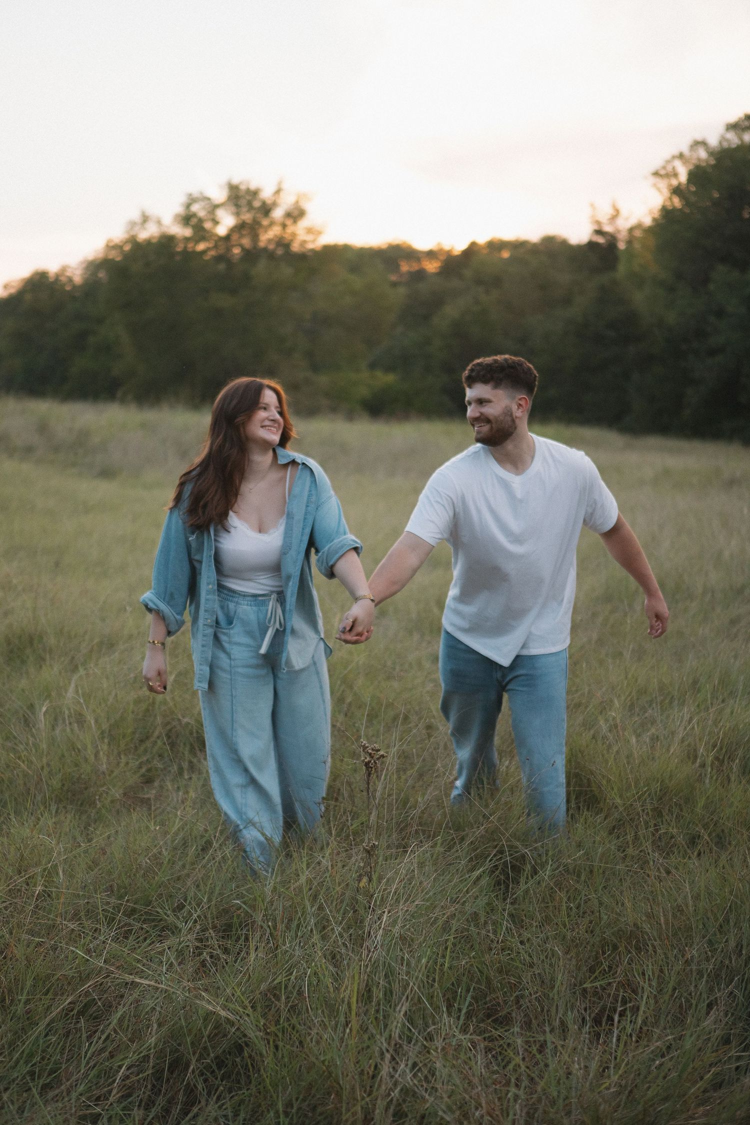 A couple walks hand in hand through a grassy meadow at sunset wearing coordinating blue and white outfits Mckinney, Texas